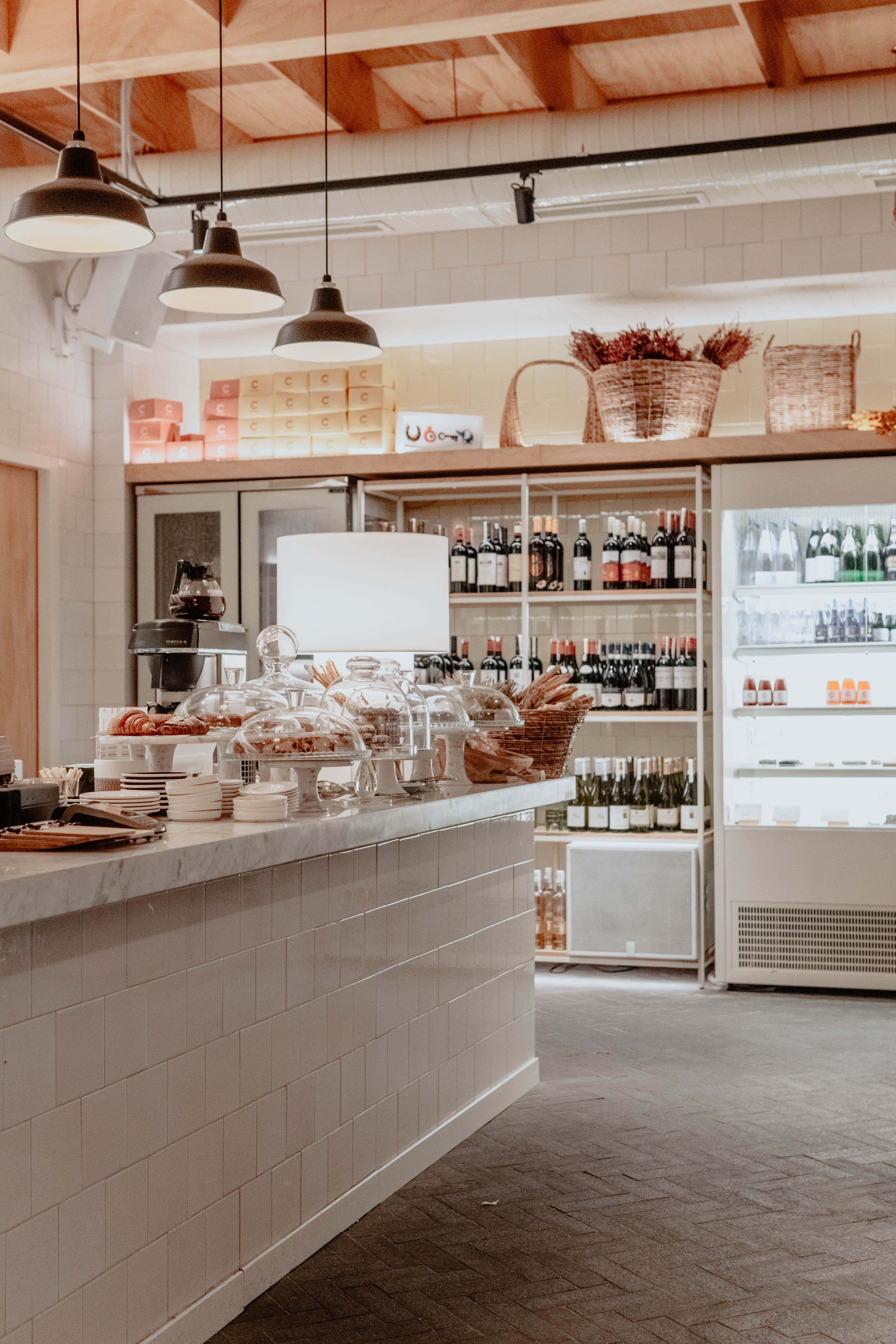 Bakery 桌布 壁紙 — 一間明亮的現代烘焙咖啡館，設有白色瓷磚檯面、玻璃罩盛載的糕點，以及懸掛吊燈下擺滿酒瓶的貨架。