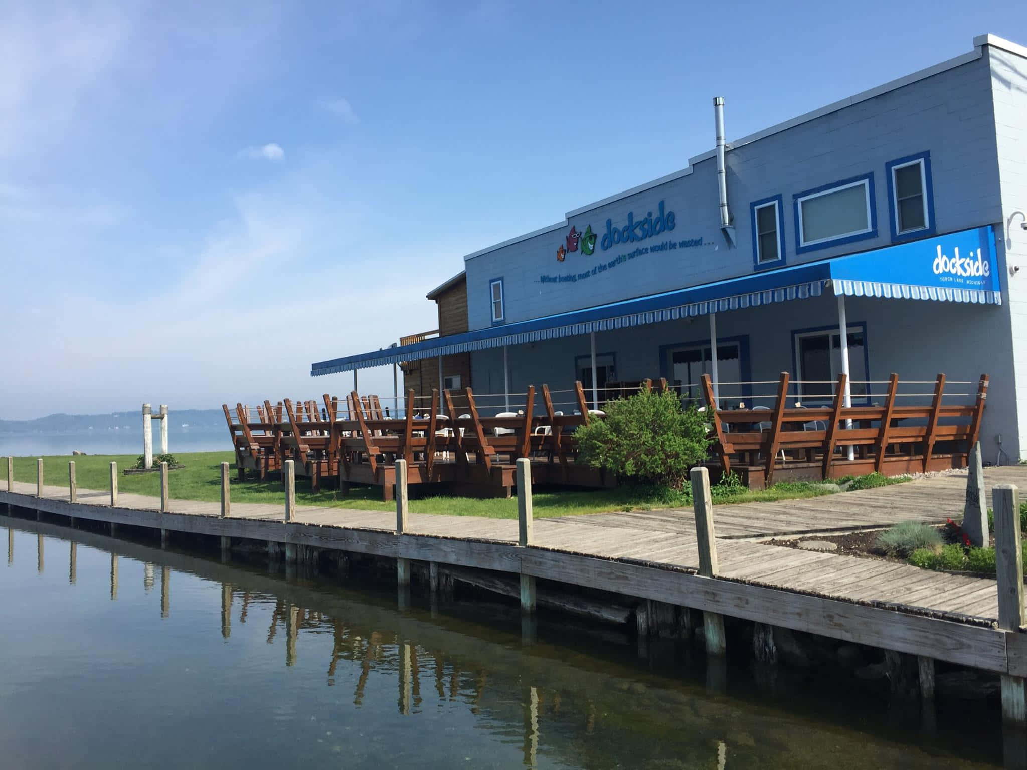 Torch Lake 桌布 壁紙 — 一間明亮的藍白色碼頭餐廳，設有木製戶外座位，可俯瞰晴朗天空下的平靜水面。