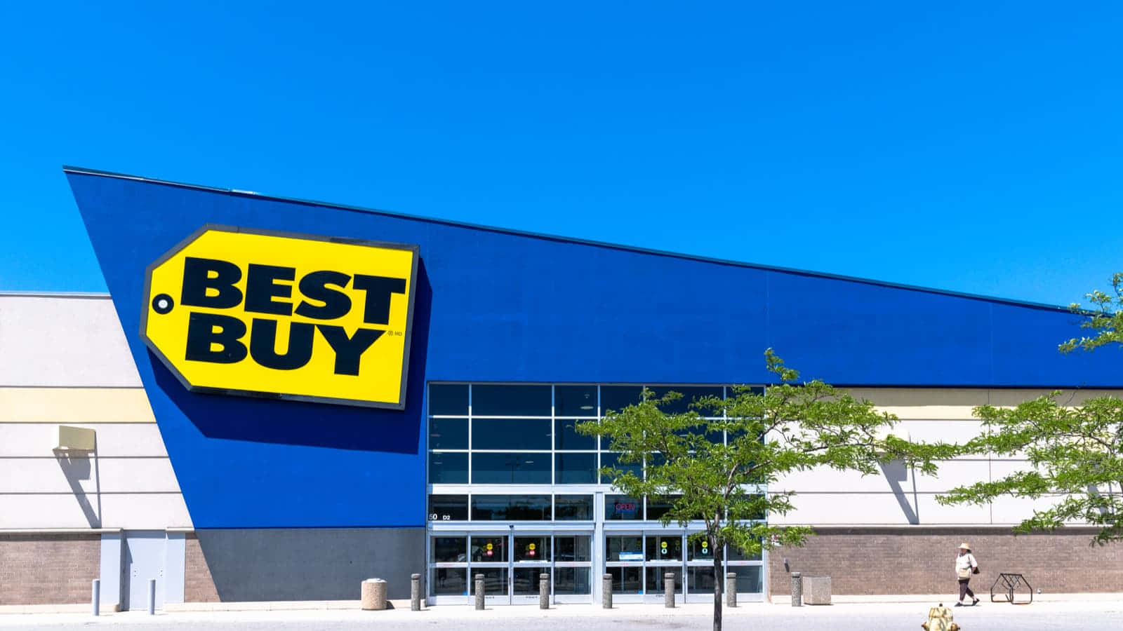 Best Buy 桌布 壁紙 — 一間明亮的藍色Best Buy商店，有一個大型黃色標誌，在晴朗的藍天下，附近有一個人走過。