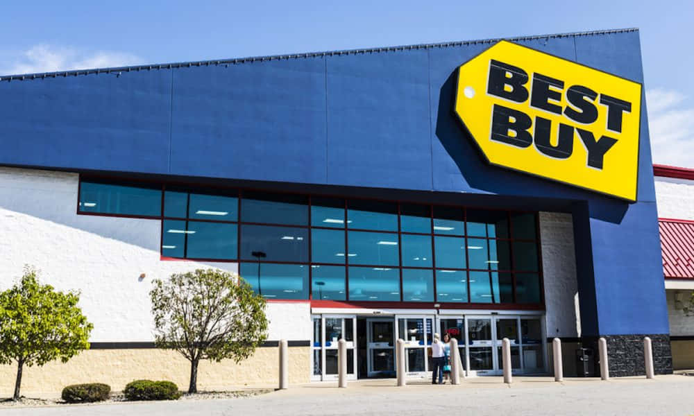 Best Buy 桌布 壁紙 — 一間明亮的藍色Best Buy商店，帶有大型黃色標籤標誌，在晴朗的天空下，有兩個人正在進入。