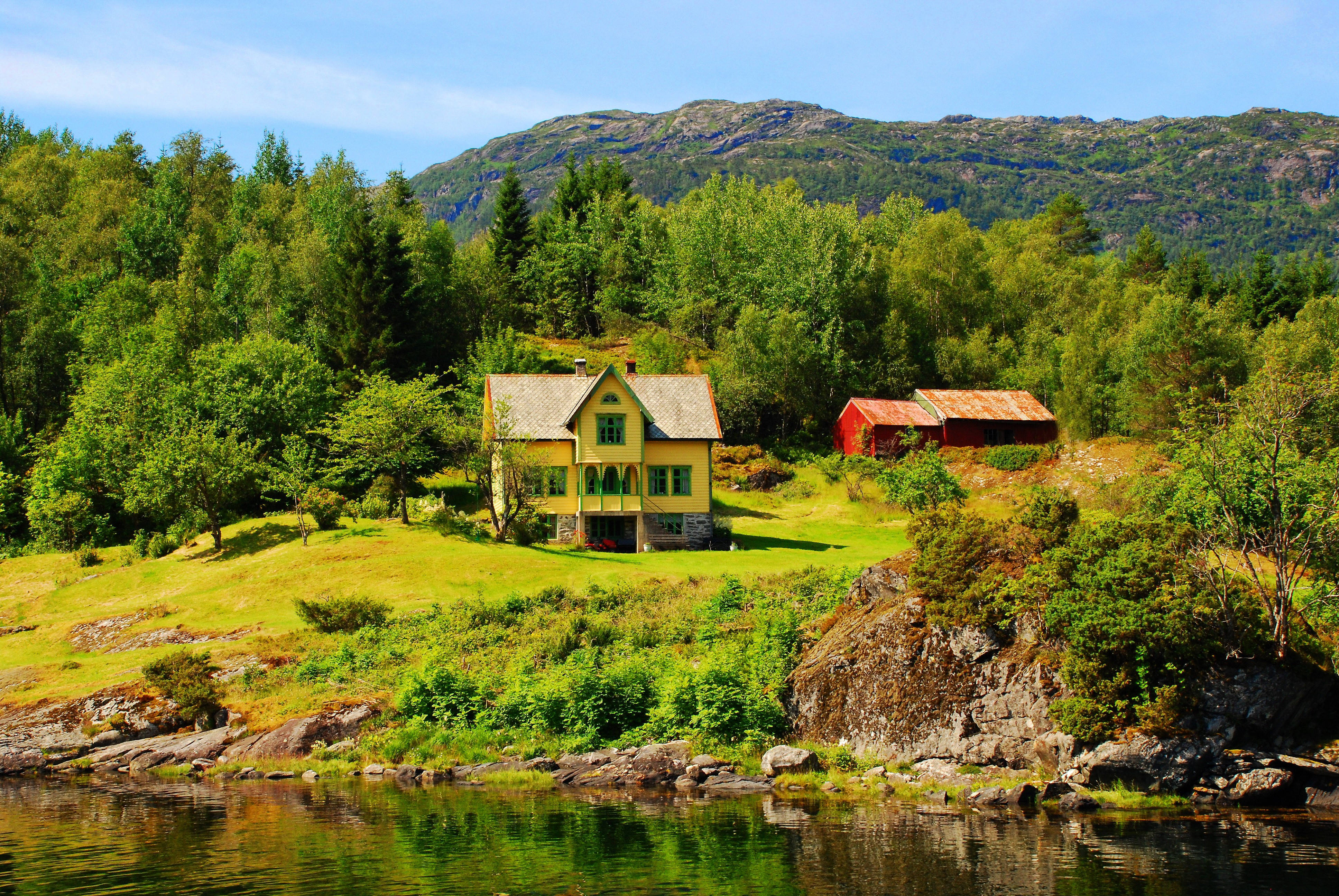 Norway 桌布 壁紙 — 一間明亮的黃色房屋和紅色穀倉坐落於草地山坡上，旁邊是平靜的水面，背景是森林覆蓋的山脈，天空清澈湛藍。