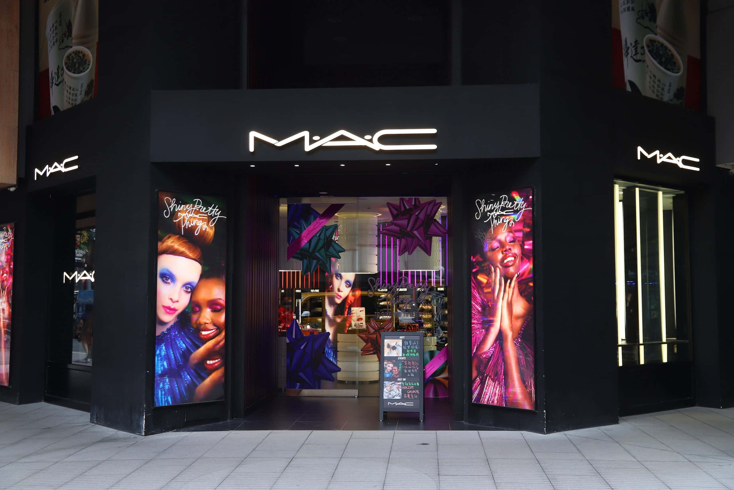 Mac Cosmetics 桌布 壁紙 — 一間時尚的黑色MAC化妝品店，帶有發光招牌和充滿活力的紫色、粉紅色和藍色宣傳海報，模特兒妝容大膽。