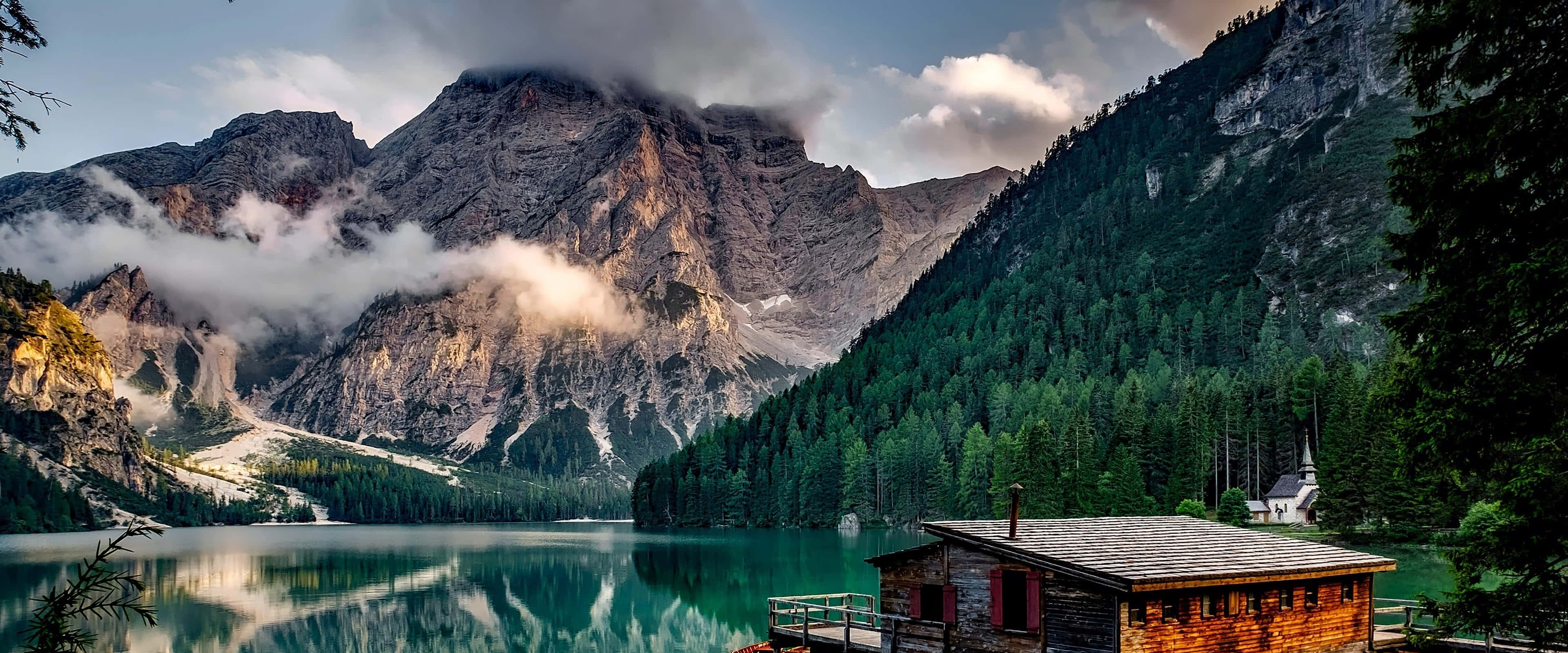 Lake 桌布 壁紙 — 一間木屋坐落在一個綠松石色的湖邊，湖面映照著高聳的山脈，山脈部分被雲遮蓋，正值日落時分。