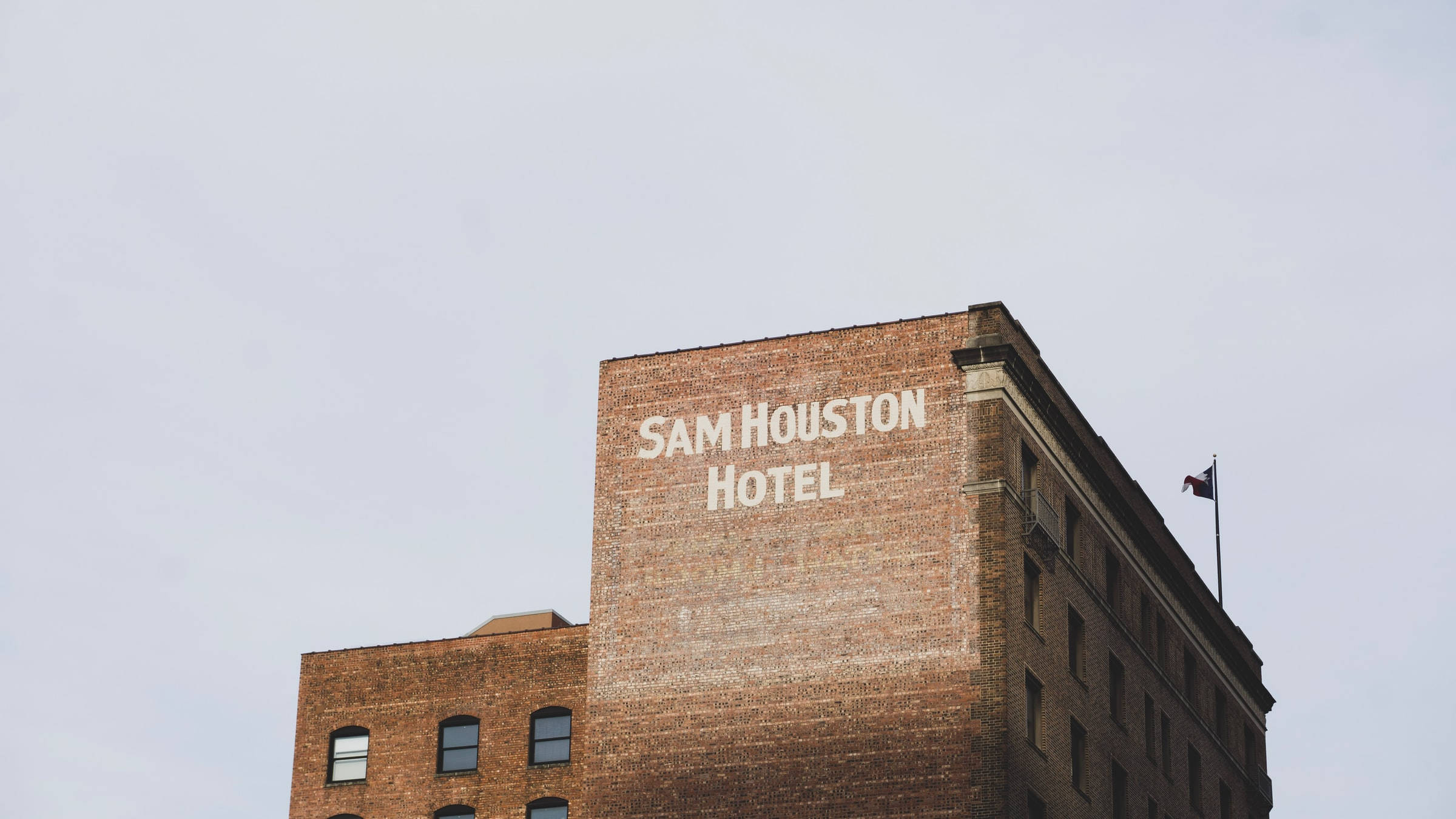 Houston 桌布 壁紙 — 一間歷史悠久的磚砌飯店，側面塗有「SAM HOUSTON HOTEL」字樣，天空多雲。