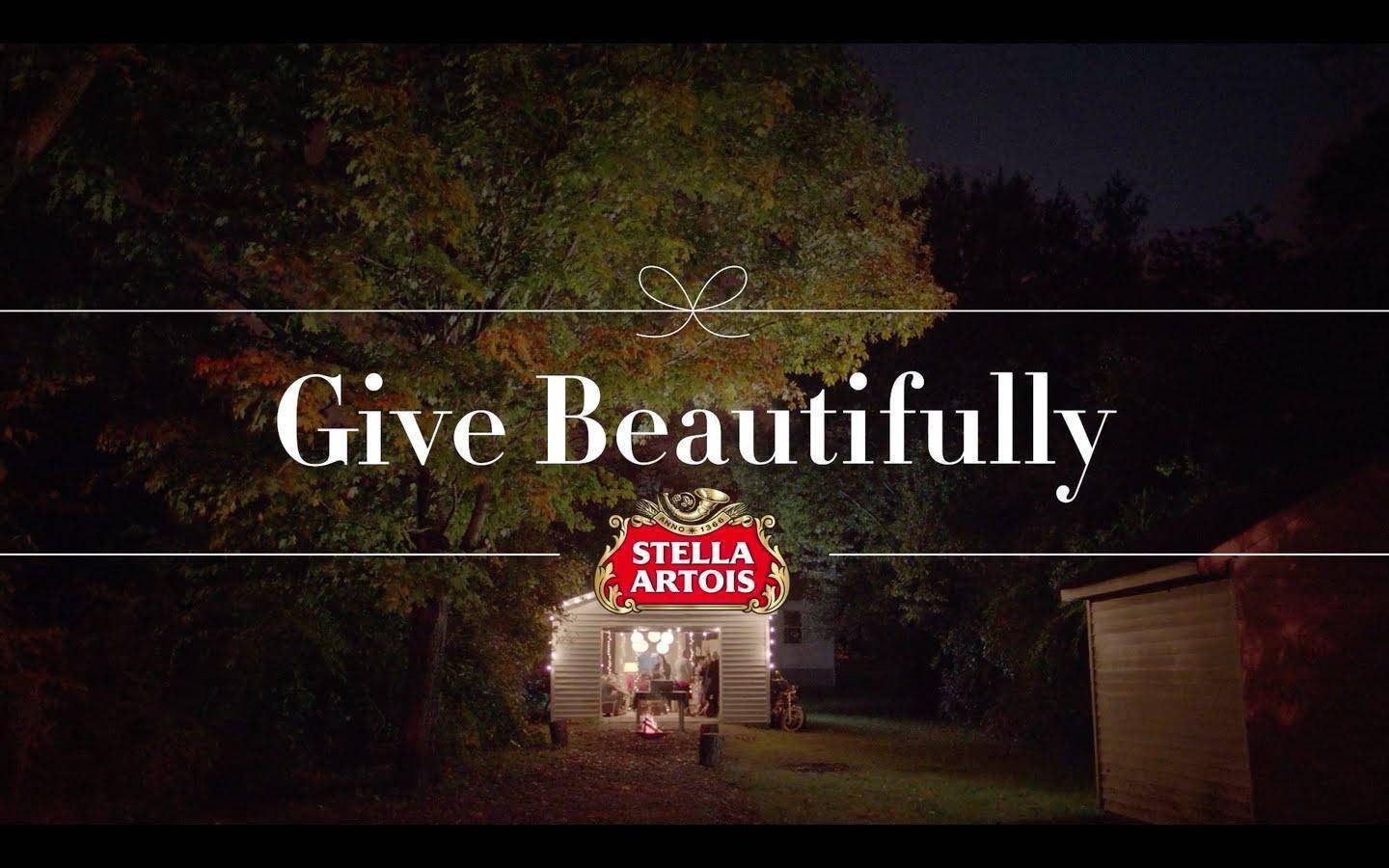 Drink 桌布 壁紙 — 一間溫暖照亮的木屋，坐落在秋夜樹下，帶有「Give Beautifully」文字和Stella Artois標誌。