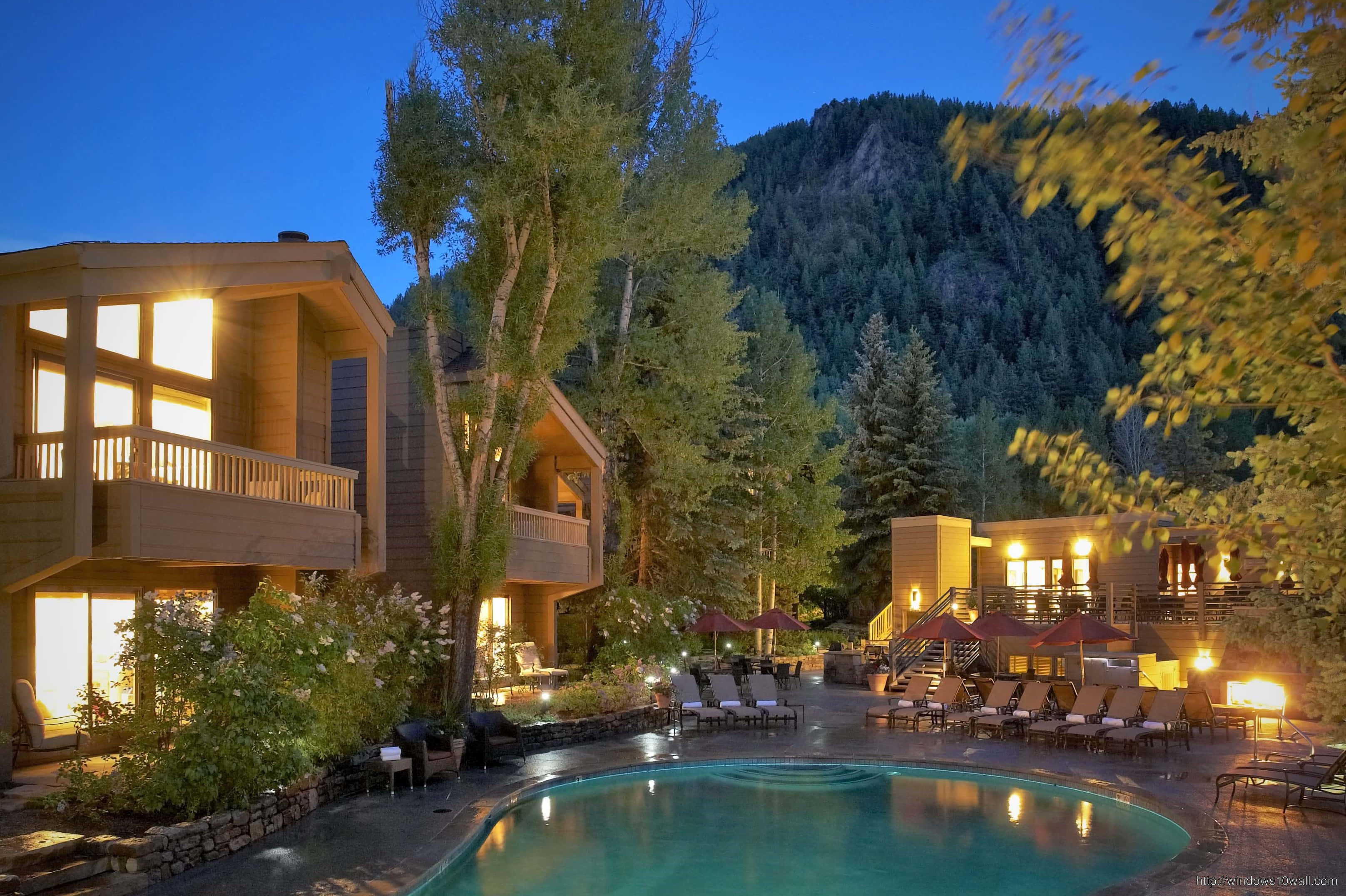 Aspen Colorado 桌布 壁紙 — 一間溫暖照明的木屋，帶有發光的游泳池，周圍環繞著樹木和山脈，在黃昏時分。