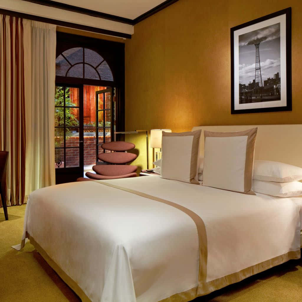 Hotel Room 桌布 壁紙 — 一間溫暖照明的飯店臥室，配有整齊鋪好的床、通往庭院的拱形玻璃門，以及芥末黃牆上裱框的城市景觀照片。