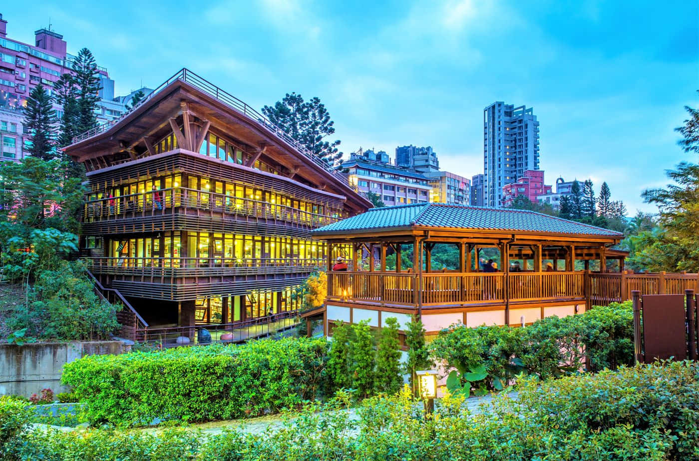Green Buildings 桌布 壁紙 — 一間溫暖的木屋，窗戶透出光芒，坐落在茂密的綠植中，背景是暮色中的城市天際線。