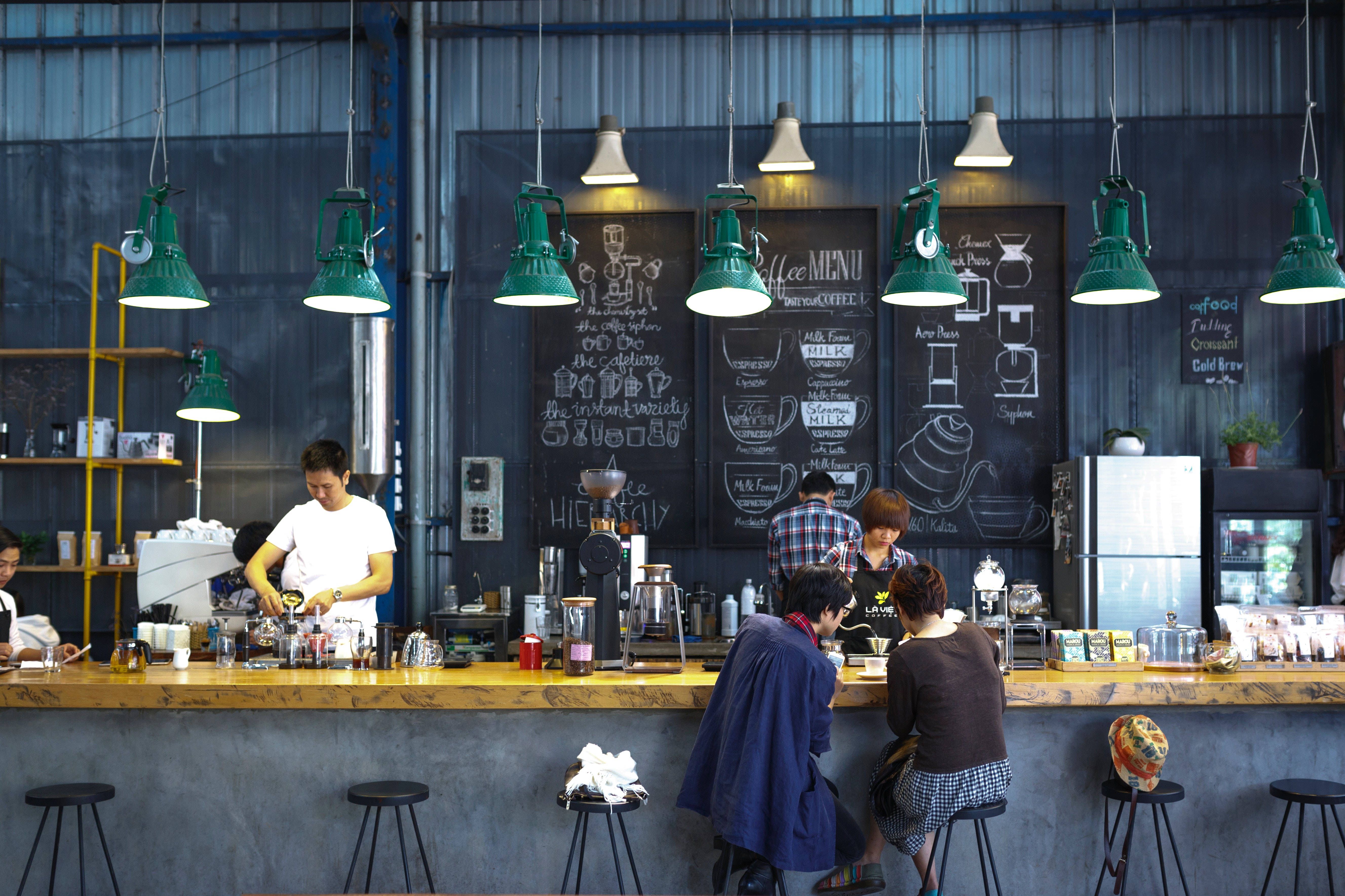 Coffee Shop 桌布 壁紙 — 一間熱鬧的工業風格咖啡店，配有綠色吊燈和黑板菜單，顧客在櫃檯前享用飲品。