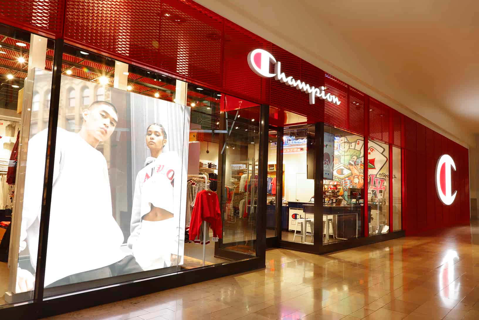Champion 桌布 壁紙 — 一間燈光明亮的Champion商店，紅色牆壁和大玻璃窗，在購物中心內展示運動服裝和人體模型。