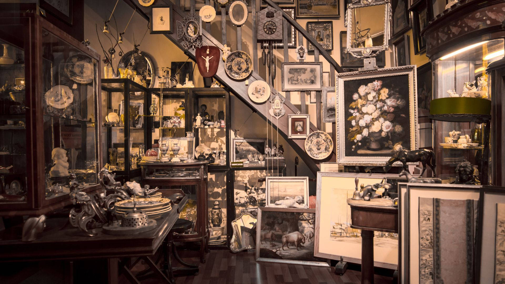 antique 桌布 壁紙 — 一間燈光溫暖的古董店，中央樓梯周圍擺滿玻璃陳列櫃、復古畫作和華麗裝飾。