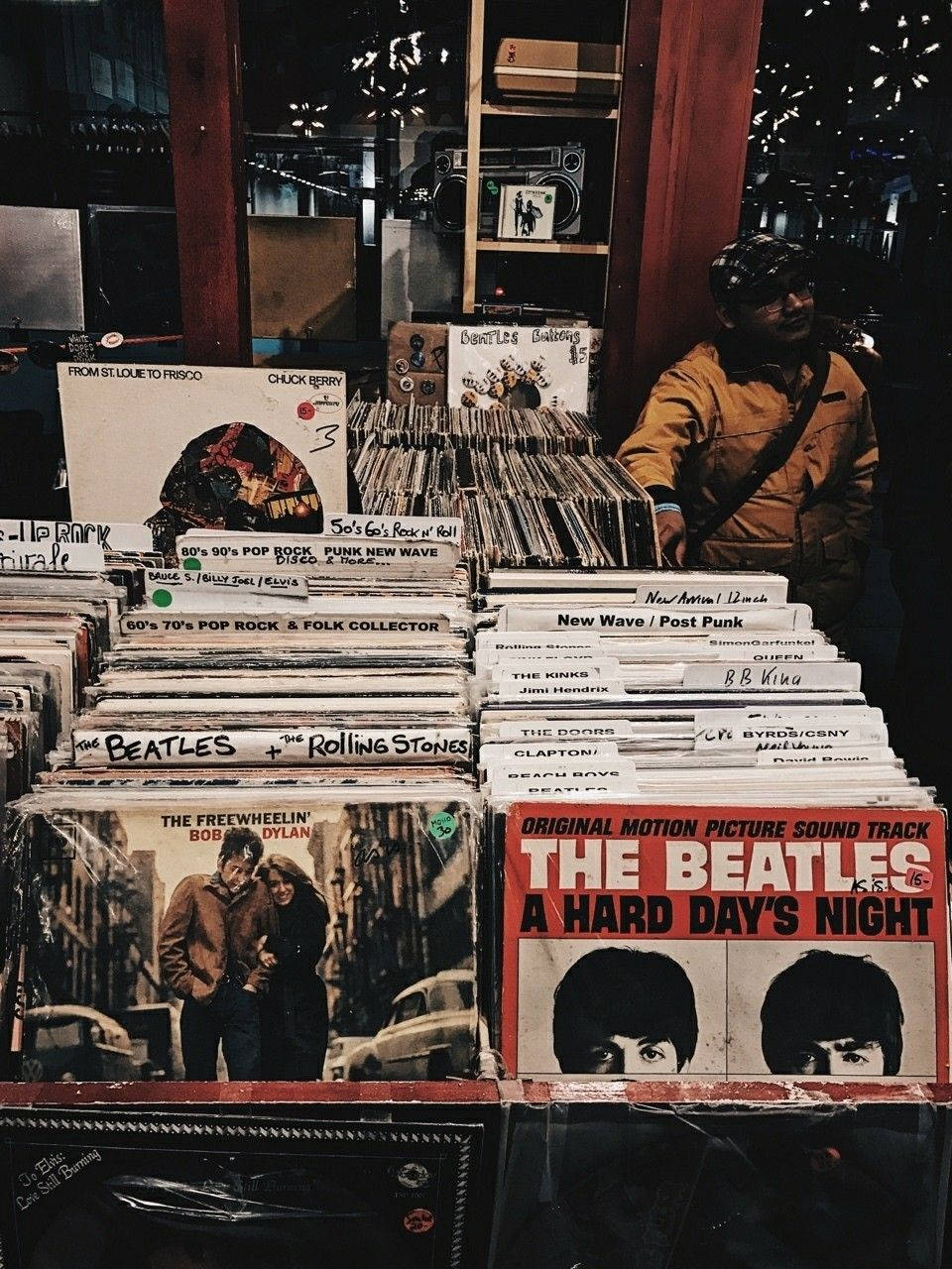 vintage 90s aesthetic 桌布 壁紙 — 一間燈光溫暖的復古唱片店展示著一堆經典黑膠唱片，背景中有一人在瀏覽。