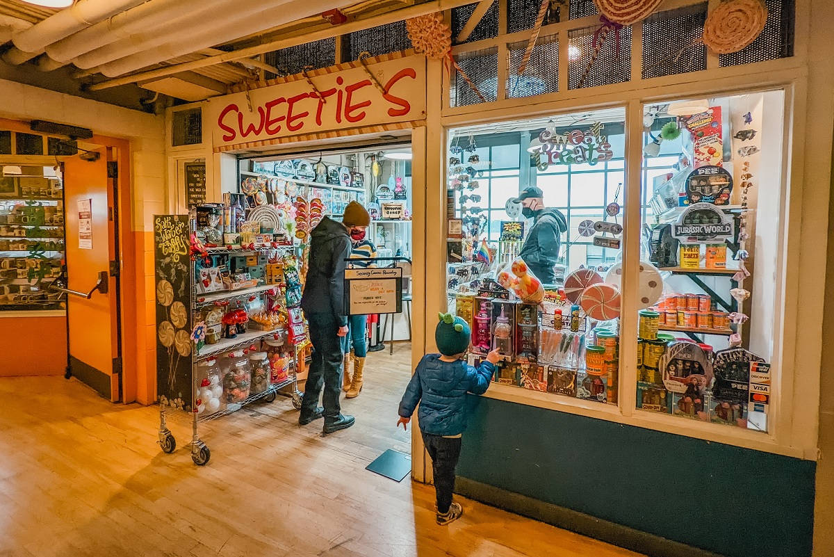 Pike Place Market 桌布 壁紙 — 一間燈光溫暖的糖果店，一個戴綠色帽子的孩子透過玻璃窗凝視著色彩繽紛的糖果，而大人們在店內購物。