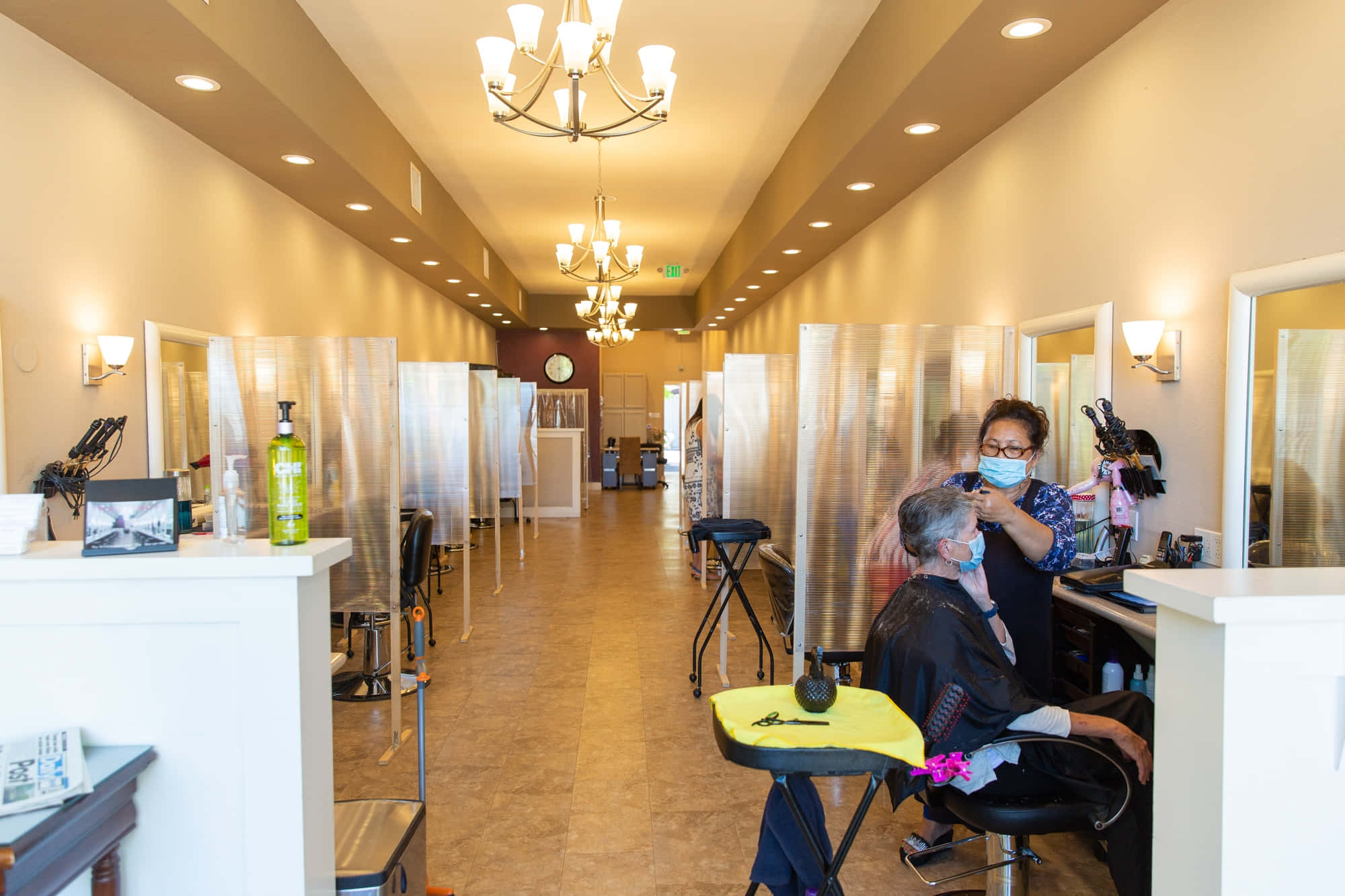 Hair Salon 桌布 壁紙 — 一間燈光溫暖的髮廊，造型師戴著口罩，在透明塑膠隔板後面為客人剪髮。