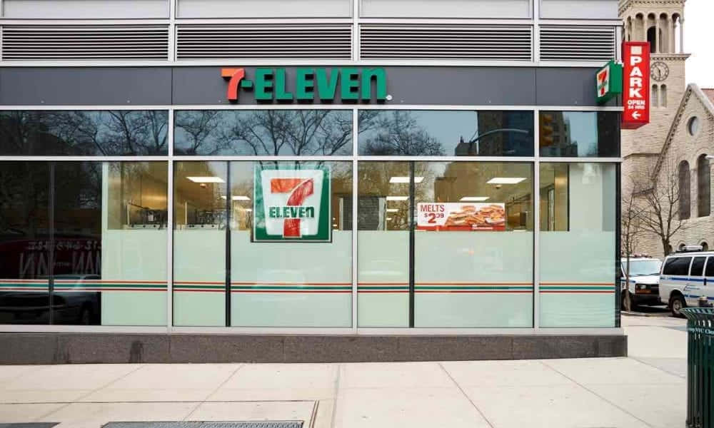 7-ELEVEN 桌布 壁紙 — 一間現代化的7-Eleven商店，配有玻璃窗和綠色招牌，矗立在城市人行道上，旁邊是一座歷史悠久的石頭教堂。