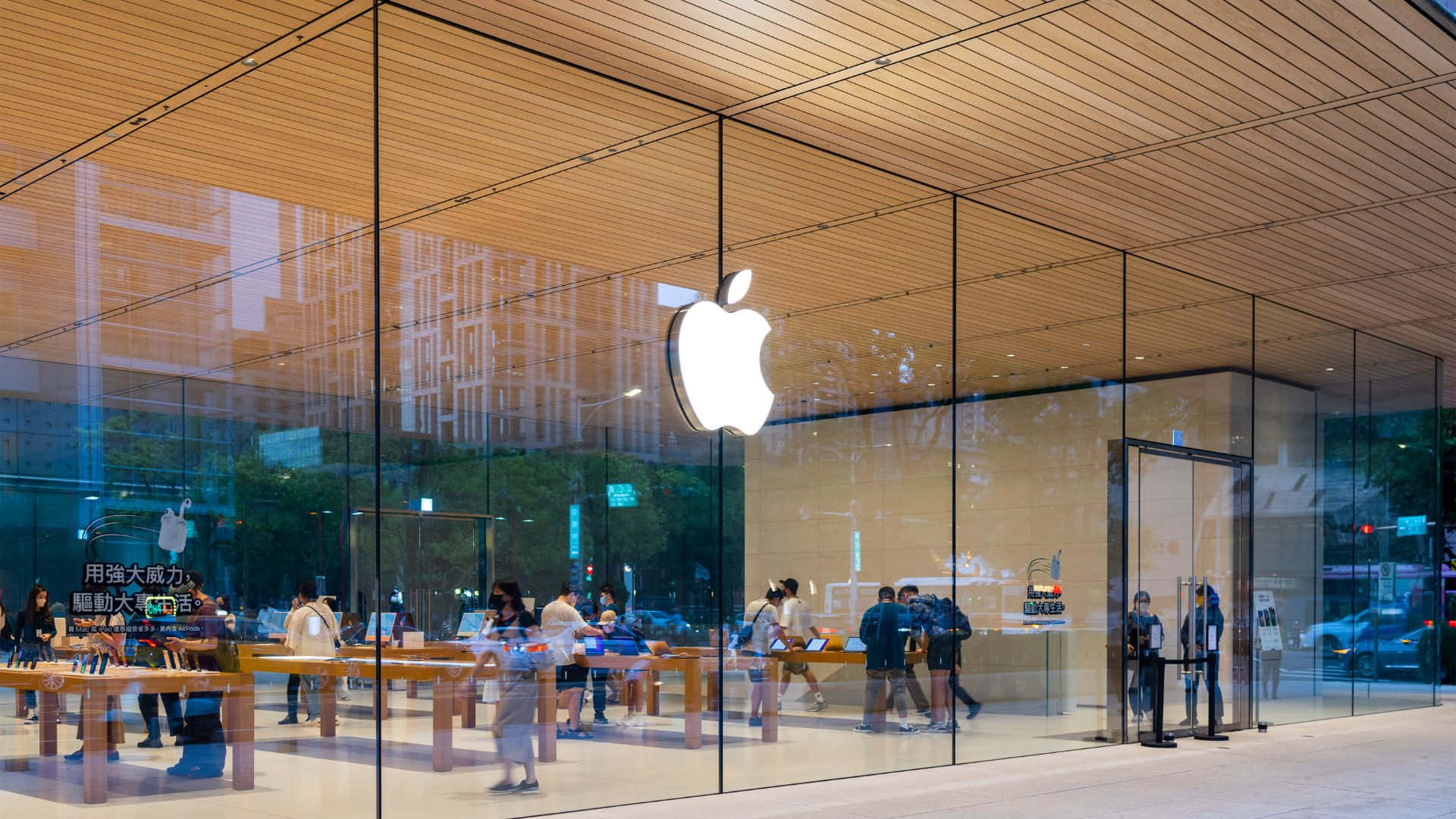 蘋果標誌 桌布 壁紙 — 一間現代化的Apple Store，設有玻璃牆、木質天花板，顧客在店內瀏覽產品。