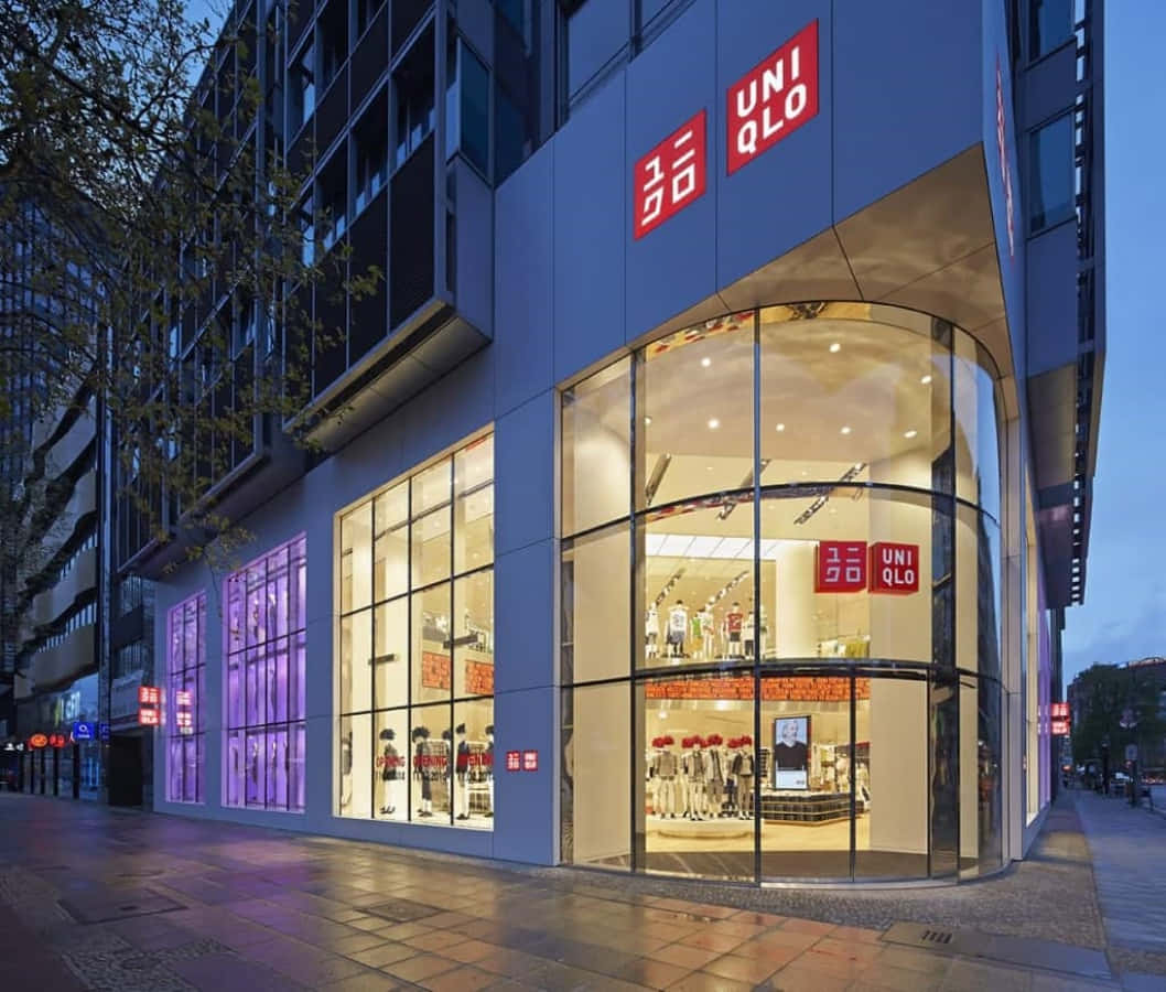 Uniqlo 桌布 壁紙 — 一間現代化的Uniqlo商店在黃昏時發出溫暖的光芒，玻璃牆展示著服裝陳列和紅色標誌，與冷色調的藍色建築外牆形成對比。
