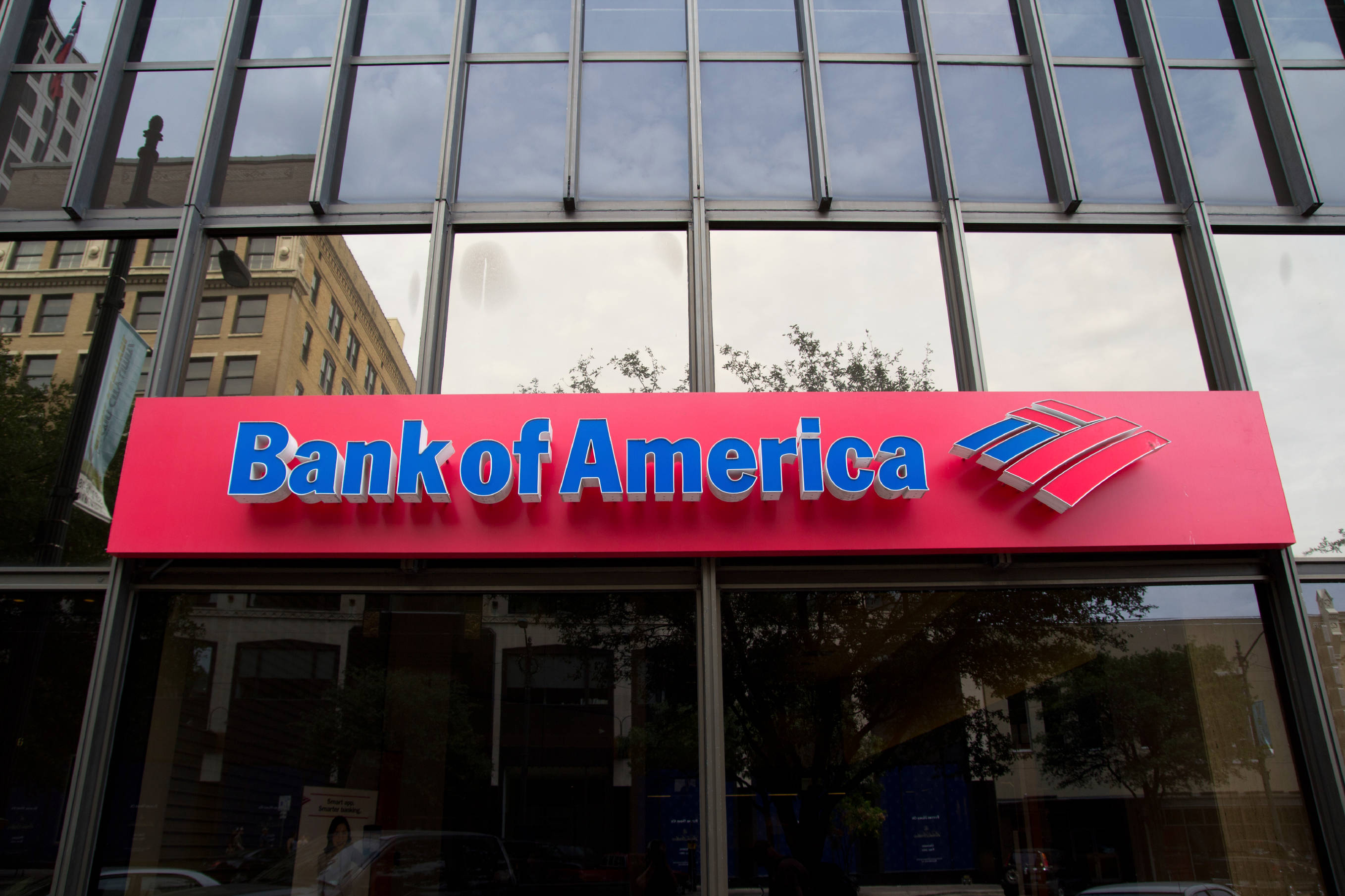 Bank 桌布 壁紙 — 一間現代化的玻璃幕牆美國銀行分行，帶有醒目的紅色標誌，映照著多雲的天空和周圍的建築物。