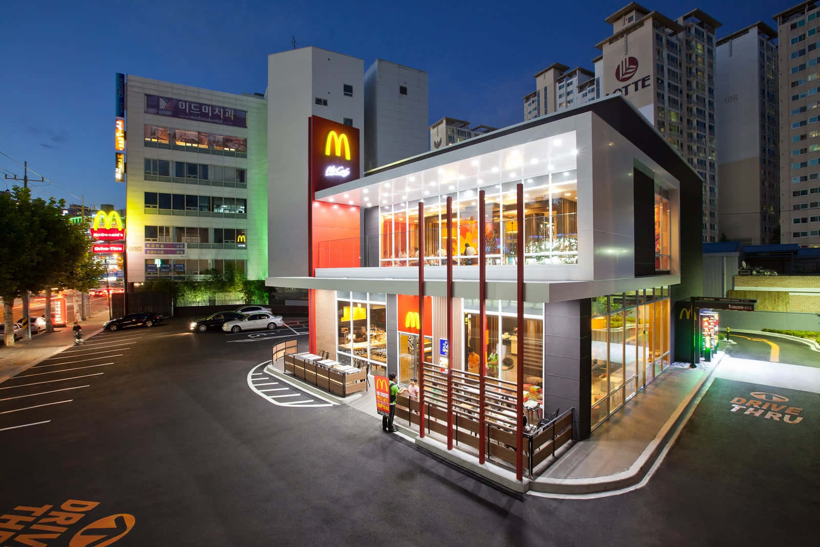 麥當勞 桌布 壁紙 — 一間現代化的麥當勞餐廳，在黃昏時分亮起燈光，窗戶閃爍，設有得來速車道，位於繁華的都市環境中。