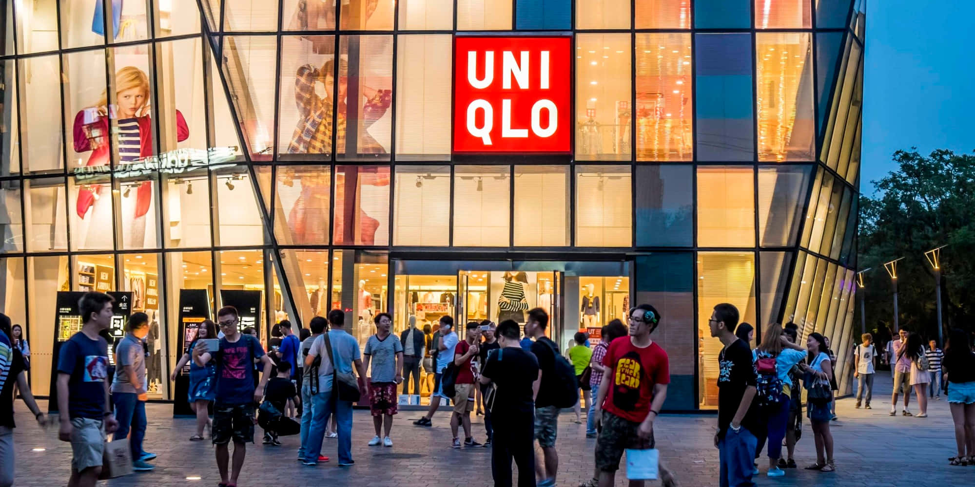 Uniqlo 桌布 壁紙 — 一間現代玻璃幕牆的Uniqlo店鋪在黃昏時溫暖地發光，人群聚集在燈火通明的入口外。