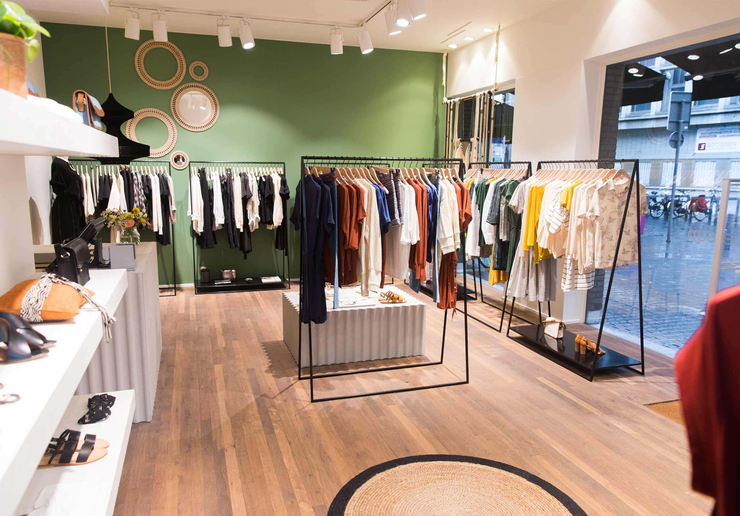 Clothes 桌布 壁紙 — 一間現代精品店，鋪有木地板、綠色牆壁，在明亮的吊燈下展示色彩繽紛的衣物架。