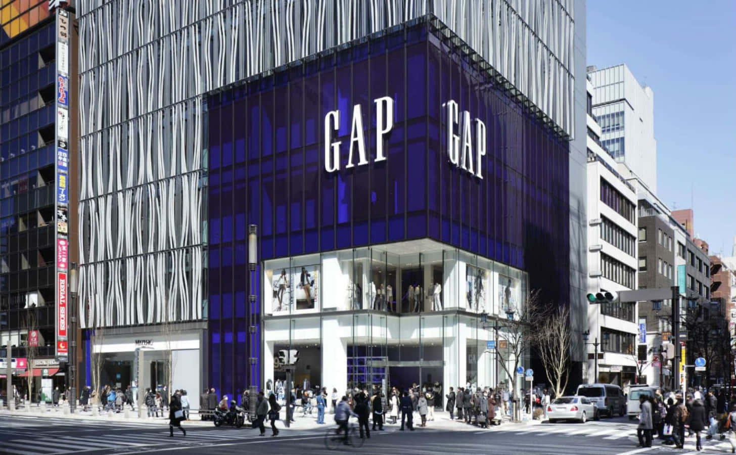 間隙 桌布 壁紙 — 一間現代藍色玻璃GAP商店，帶有白色標誌，在晴朗的白天矗立於繁忙的都市街道上，街道上滿是行人和車輛。