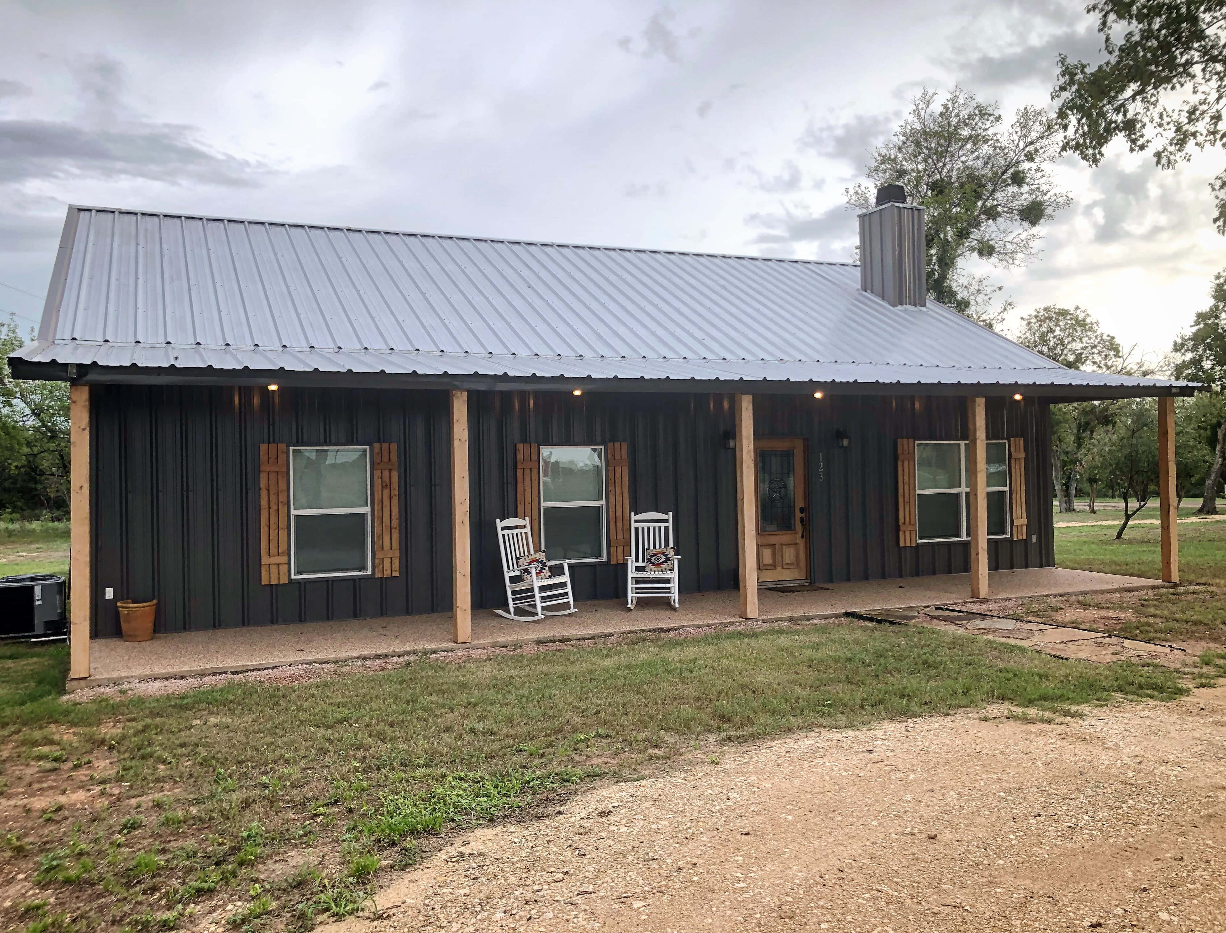 房屋 桌布 壁紙 — 一間現代鄉村風格的小屋，採用深色壁板和金屬屋頂，設有帶頂棚的門廊，在陰天之下擺放著兩張白色搖椅。