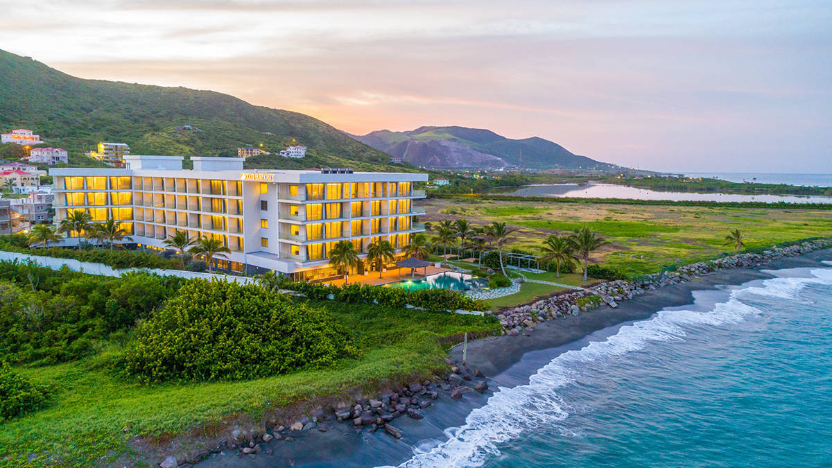 Hotel 桌布 壁紙 — 一間現代酒店在日落時分溫暖地發光，旁邊是黑色沙灘，綠松石色的海浪拍打著綠色山丘。