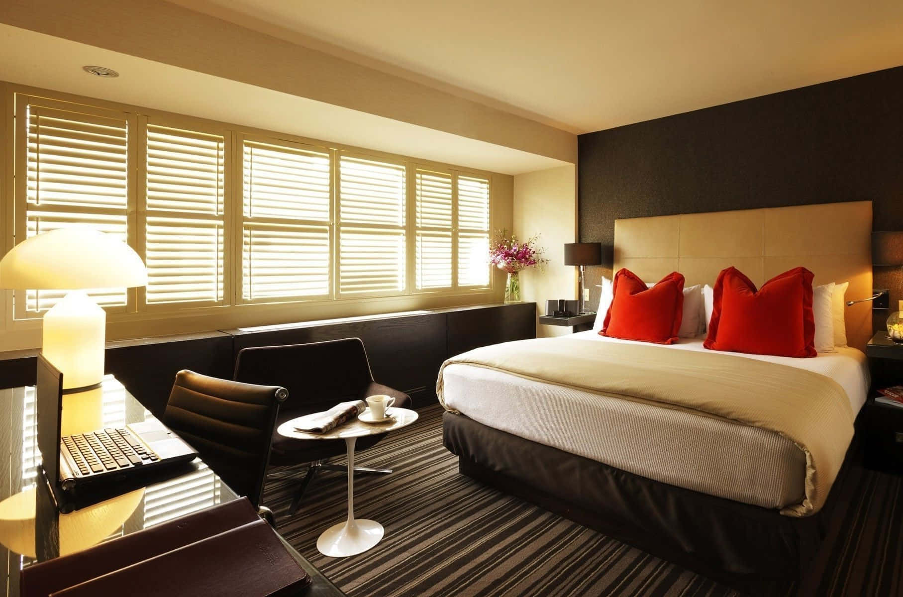 Hotel Room 桌布 壁紙 — 一間現代酒店客房，透過木百葉窗灑滿溫暖陽光，床上以鮮明的紅色枕頭作點綴。