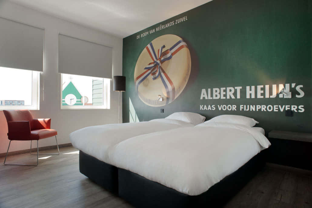 Bedroom 桌布 壁紙 — 一間現代酒店客房，配有一張大床、紅色椅子，以及一面帶有巨型起司廣告的綠色牆壁。