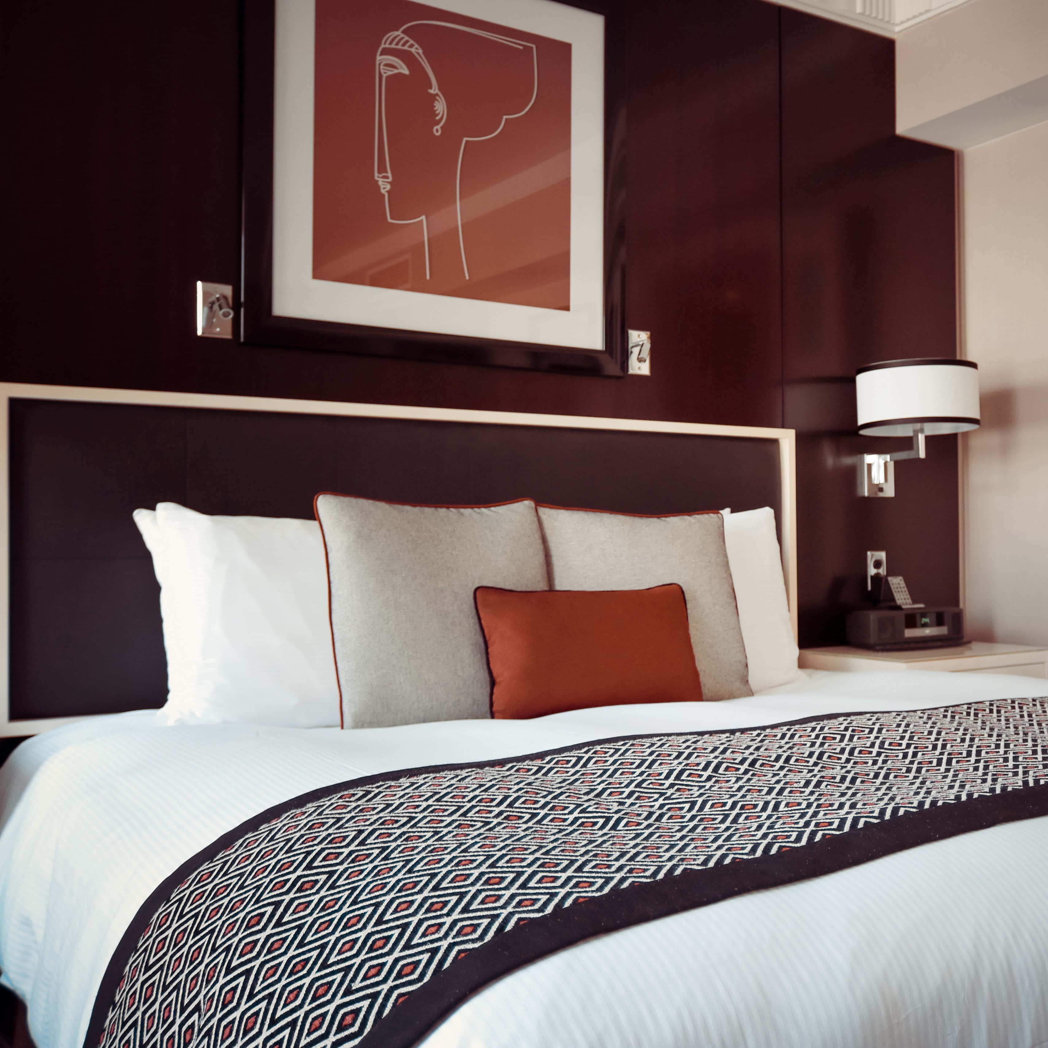 Hotel Room 桌布 壁紙 — 一間現代酒店臥室，床鋪整齊，配有幾何圖案床尾巾，深色床頭板上方掛有抽象藝術品。