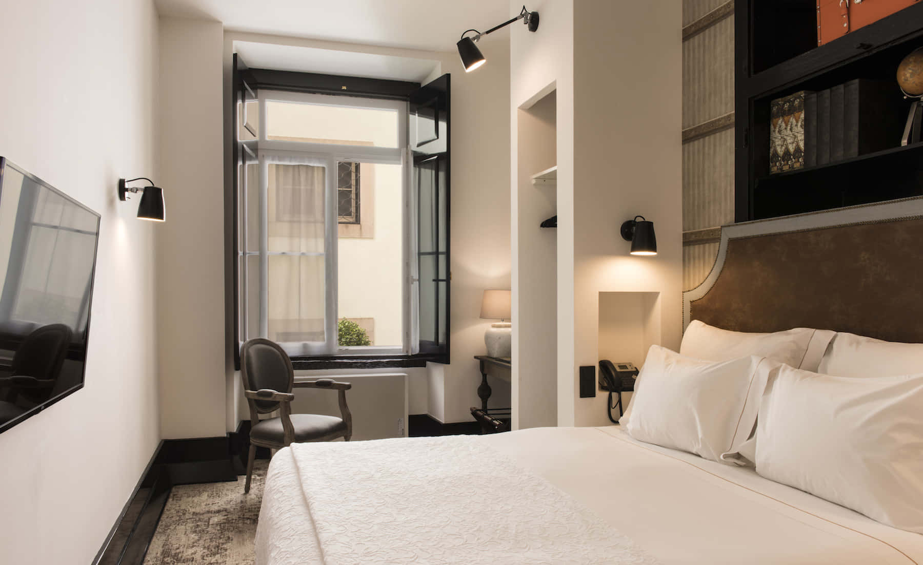 Hotel Room 桌布 壁紙 — 一間現代飯店客房，設有整齊鋪好的床、通往庭院的開放式窗戶，以及來自壁燈的溫暖環境照明。