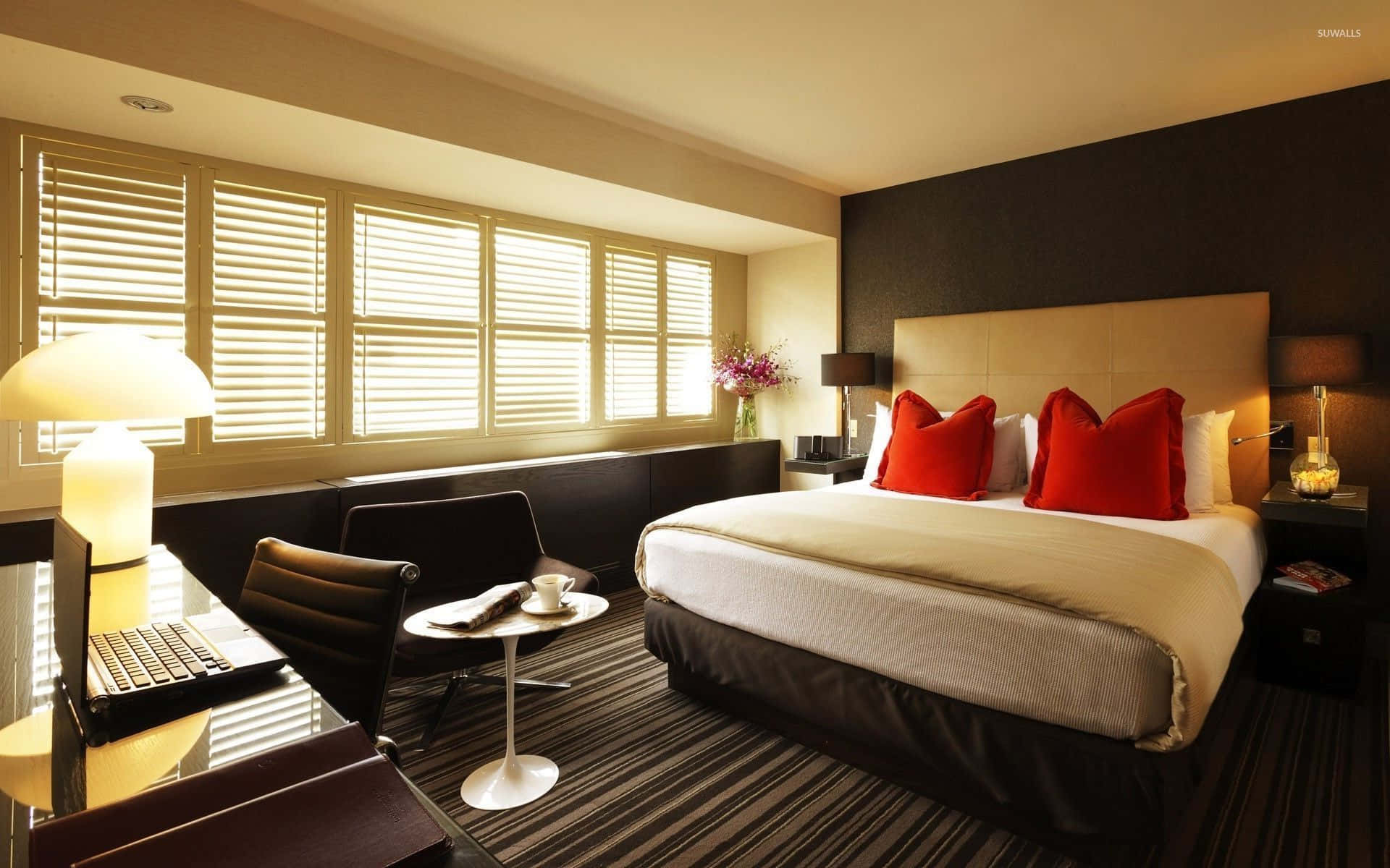 Bedroom 桌布 壁紙 — 一間現代飯店客房，透過木百葉窗灑入溫暖陽光，大床上點綴著大膽的紅色枕頭。