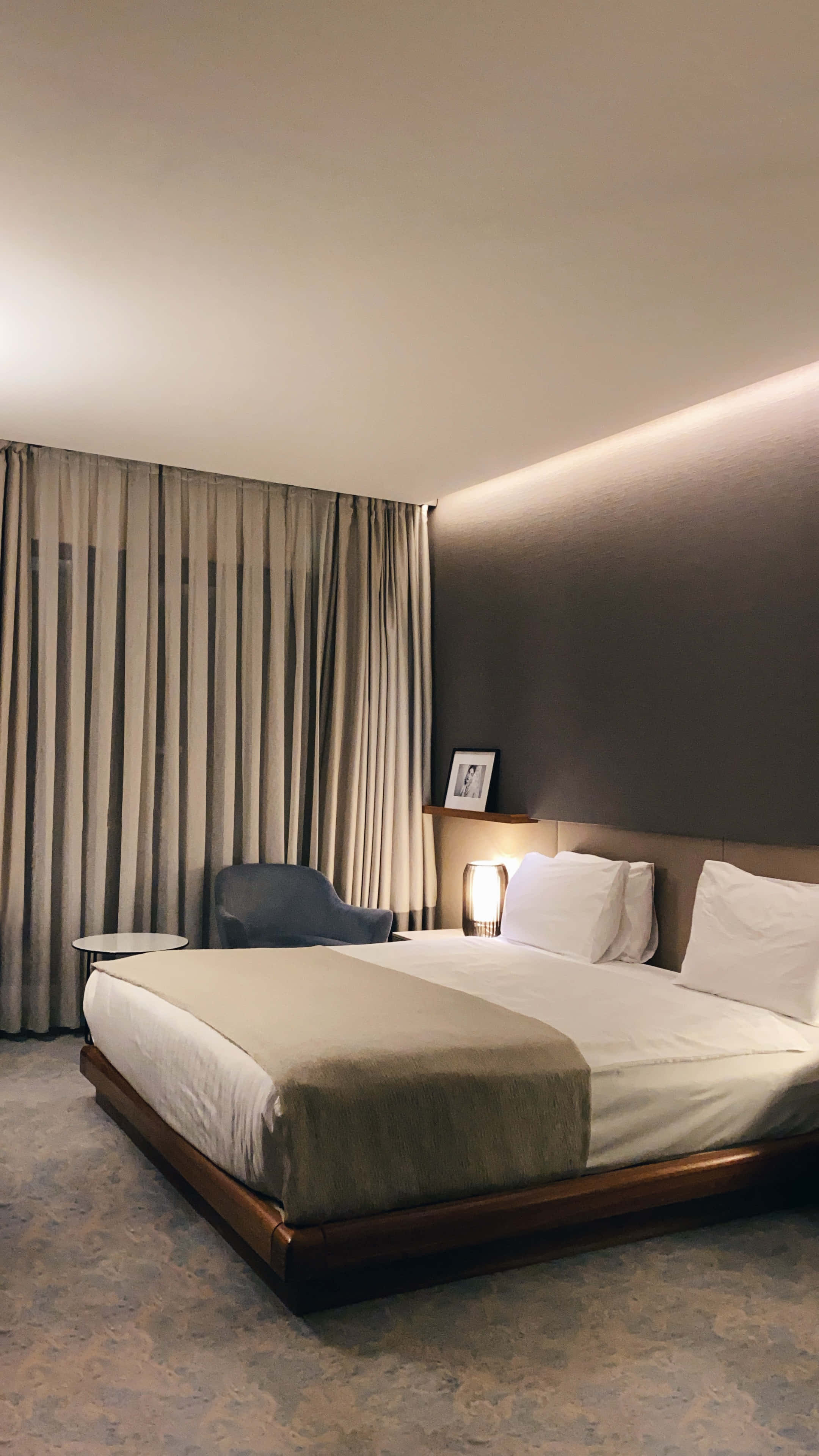 Hotel Room 桌布 壁紙 — 一間現代飯店客房，配有一張整齊鋪好的床，在溫暖的環境燈光和落地窗簾下。