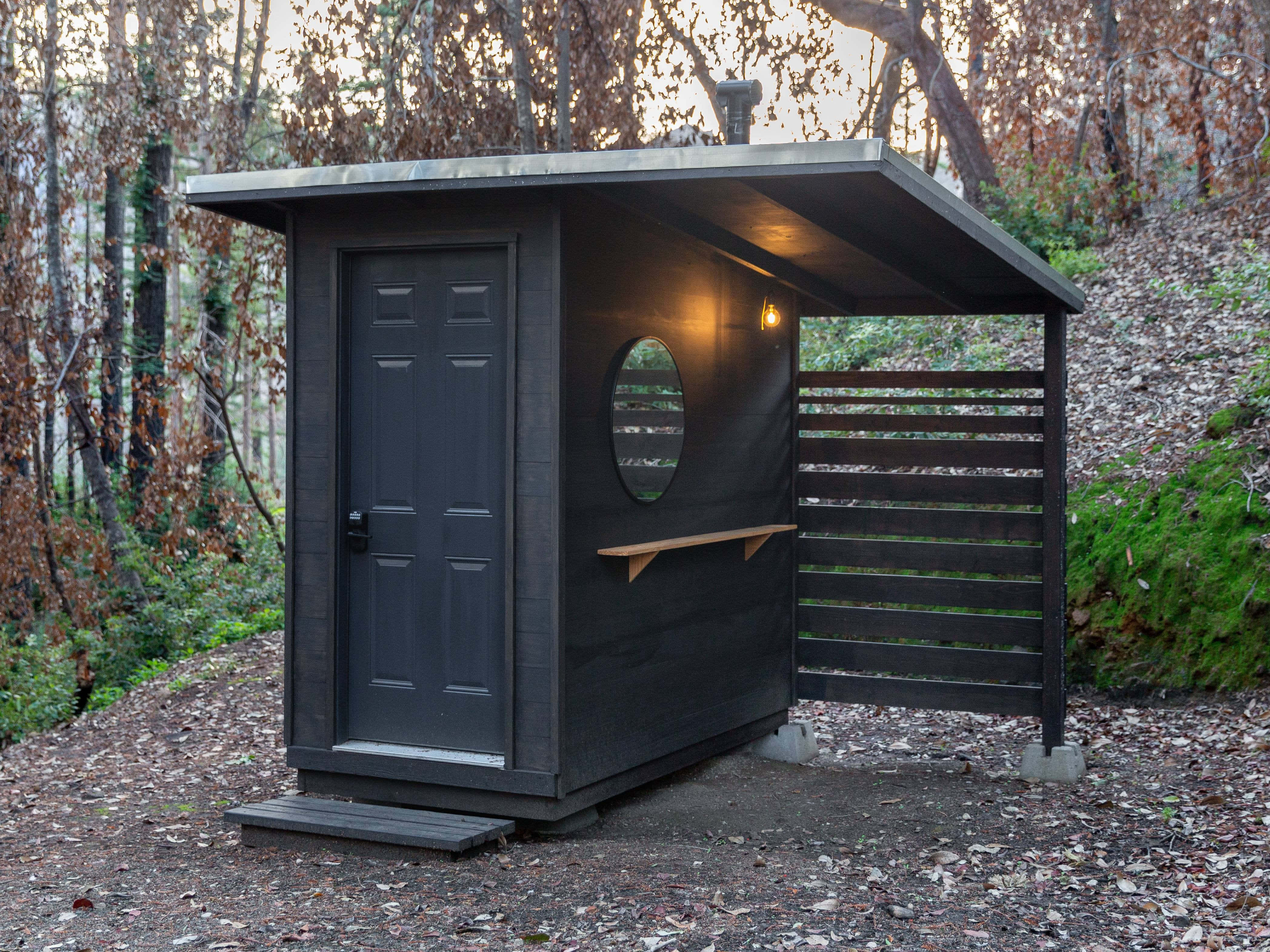 Outhouse 桌布 壁紙 — 一間現代黑色桑拿小屋，配有圓窗和木凳，坐落在黃昏的森林中。