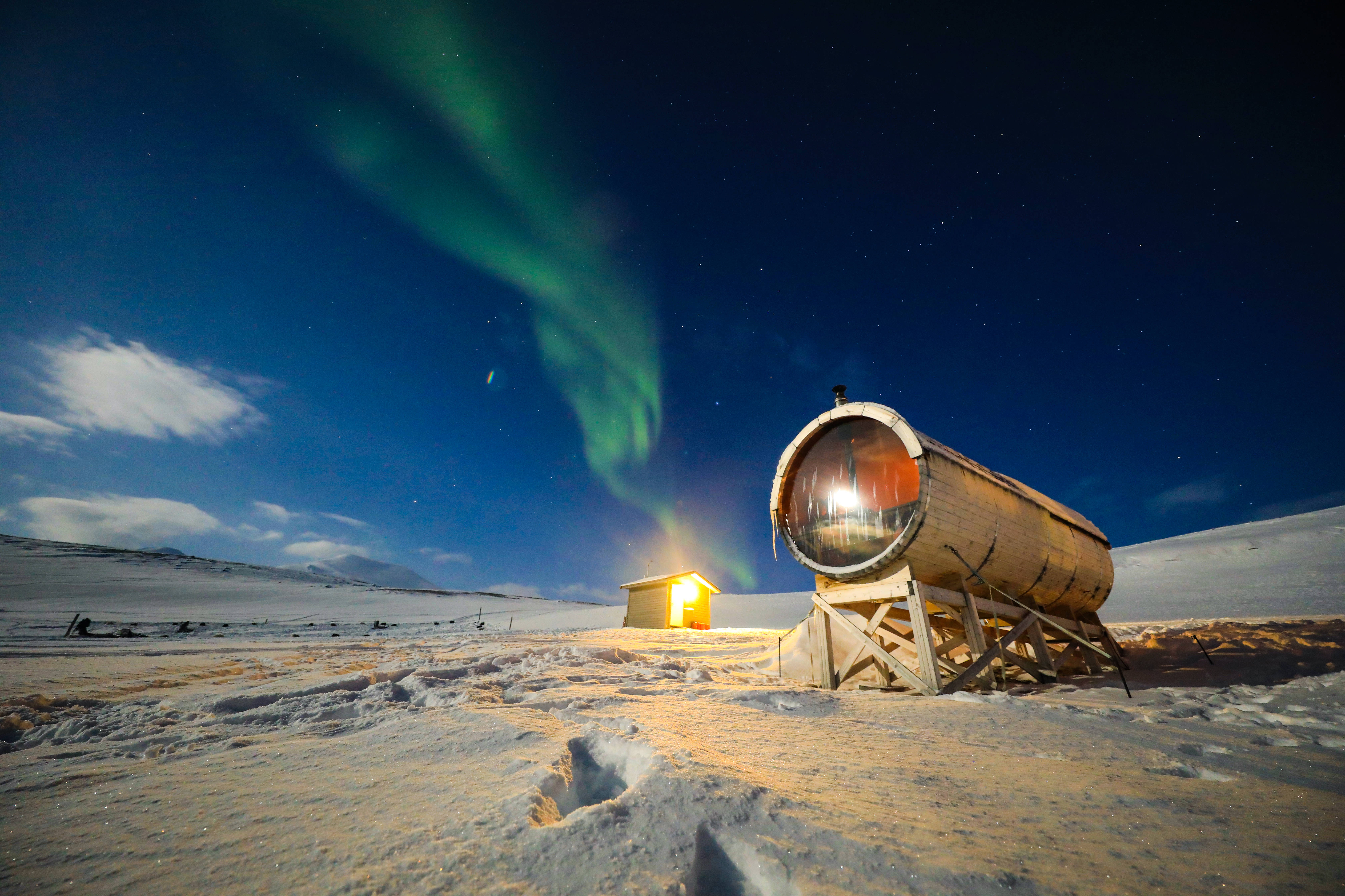 Svalbard 桌布 壁紙 — 一間發光的木製桑拿房和小屋矗立在白雪覆蓋的荒野中，翠綠的北極光在繁星點點的夜空中舞動。