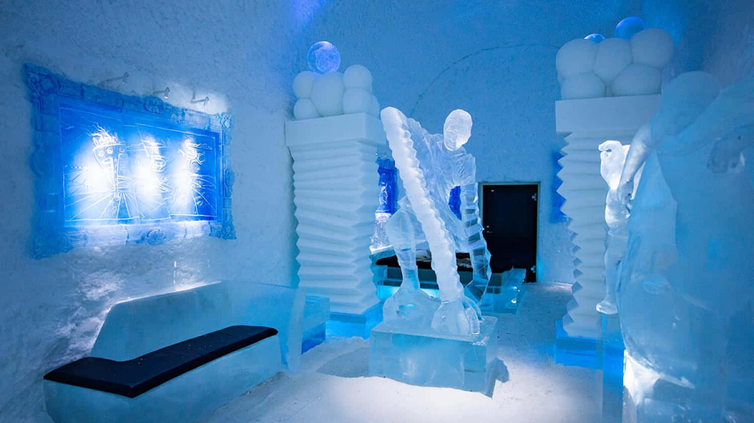 Ice Hotel 桌布 壁紙 — 一間發光的藍色冰酒店房間，設有精緻的雕塑和一個由冷光照射的帶框冰藝術品。