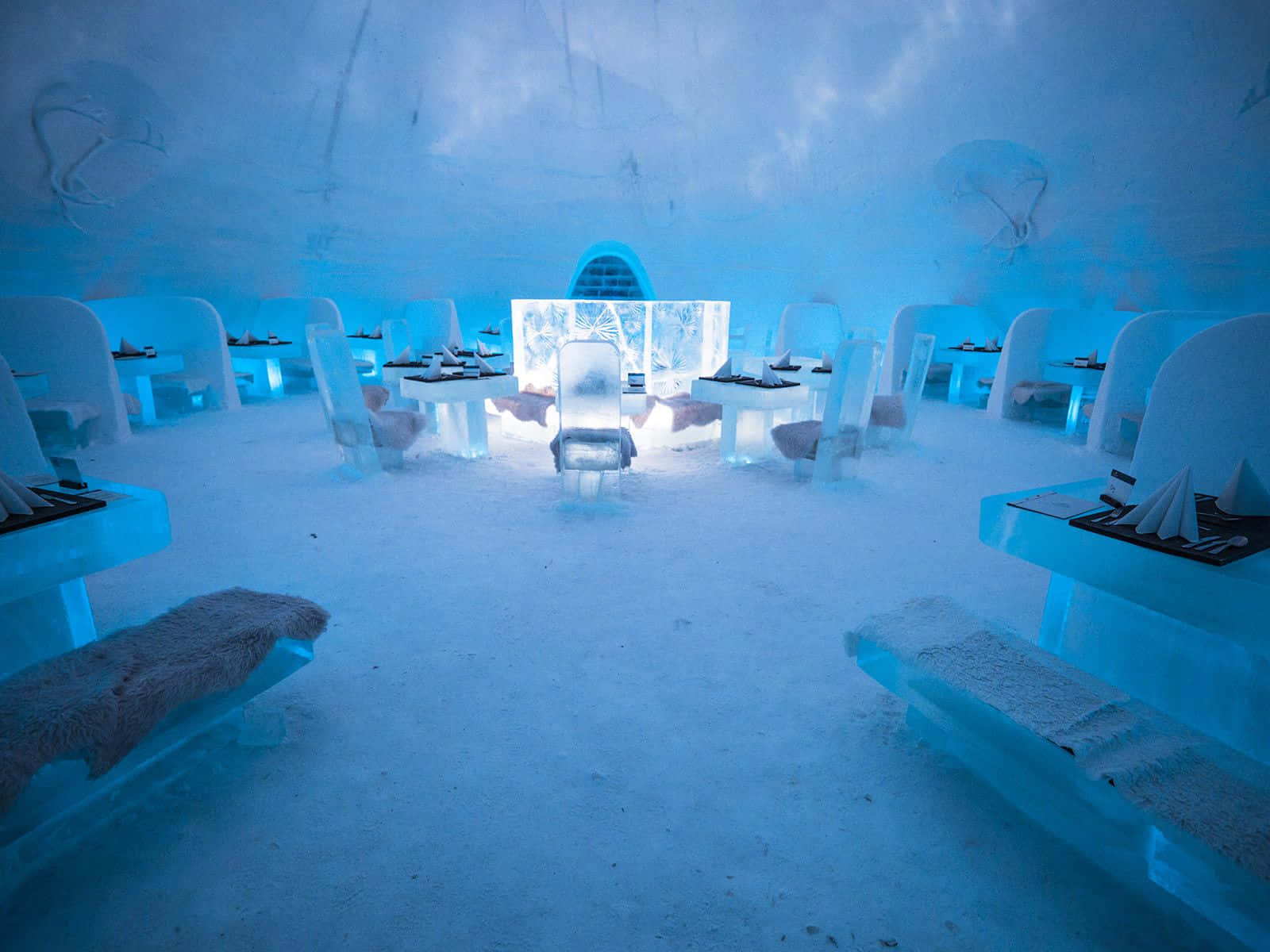 Ice Hotel 桌布 壁紙 — 一間發光的藍色冰餐廳，在彎曲的冰天花板下設有雕刻的桌子、椅子和長凳。