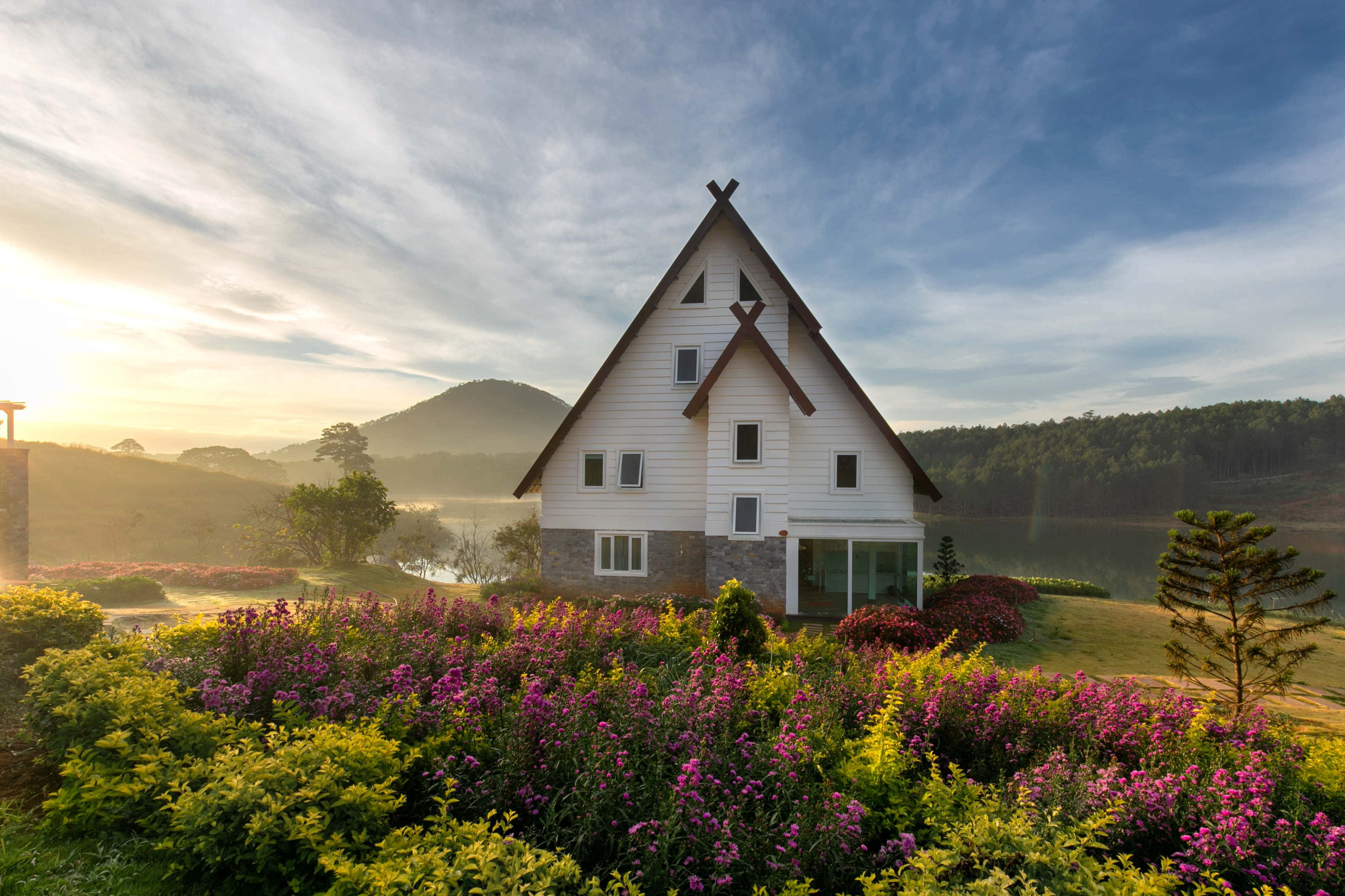 Cottage Garden 桌布 壁紙 — 一間白色A字形房屋坐落在充滿活力的紫色和黃色花朵中，背景是霧氣繚繞的山脈和湖泊，正值日出時分。