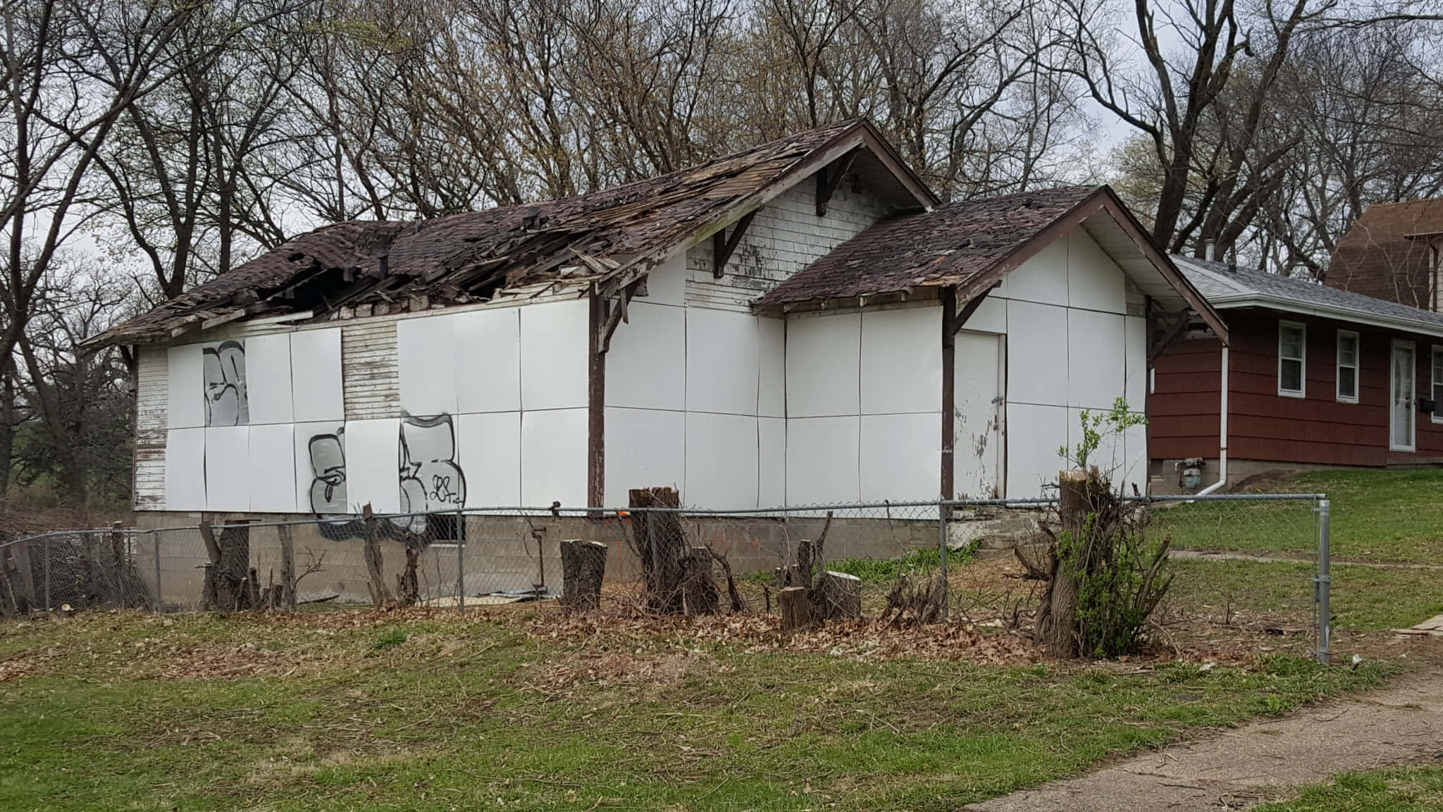 Trap House 桌布 壁紙 — 一間破舊的白屋，屋頂受損並有塗鴉，矗立在鐵絲網圍欄後的荒蕪庭院中。