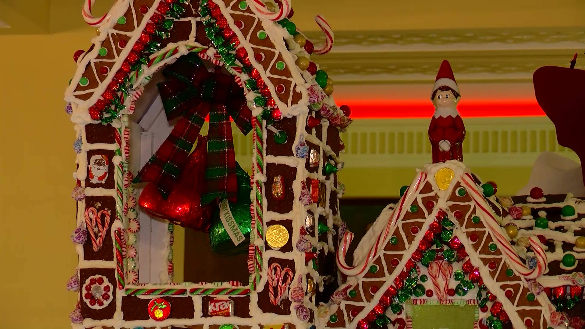 Gingerbread House 桌布 壁紙 — 一間節日薑餅屋，裝飾著拐杖糖、紅色蝴蝶結和閃閃發光的飾品，旁邊站著一個小精靈公仔。