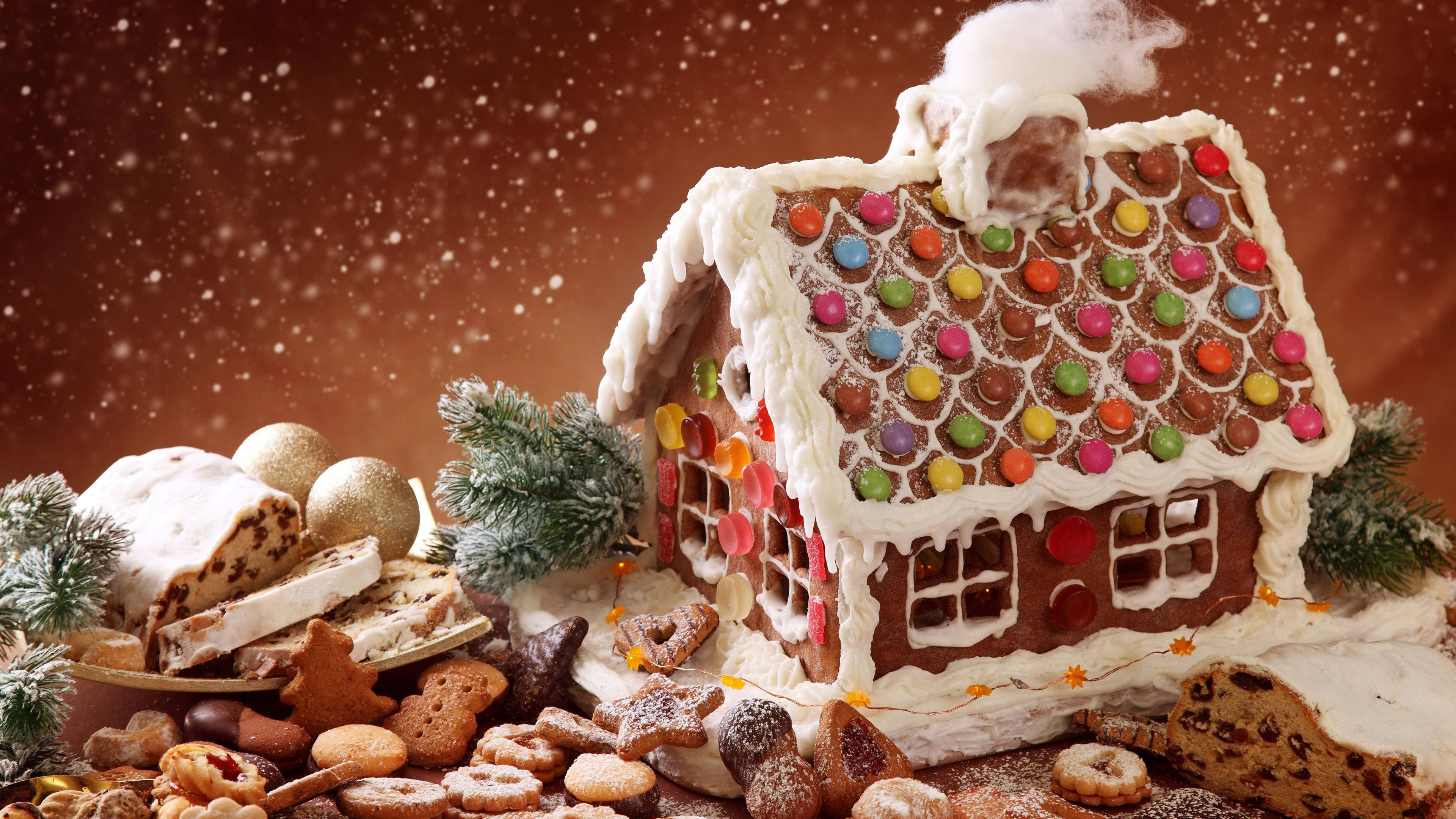 Gingerbread House 桌布 壁紙 — 一間節日薑餅屋，飾有彩色糖果和白色糖霜，坐落在白雪覆蓋的松樹枝和各種節日餅乾之間，背景是飄落的雪花。