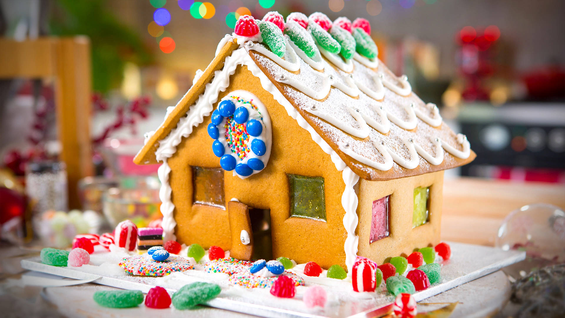 Gingerbread House 桌布 壁紙 — 一間節日薑餅屋，飾有色彩繽紛的糖果和糖粒，放置在雪白底座上，周圍環繞著節日美食。