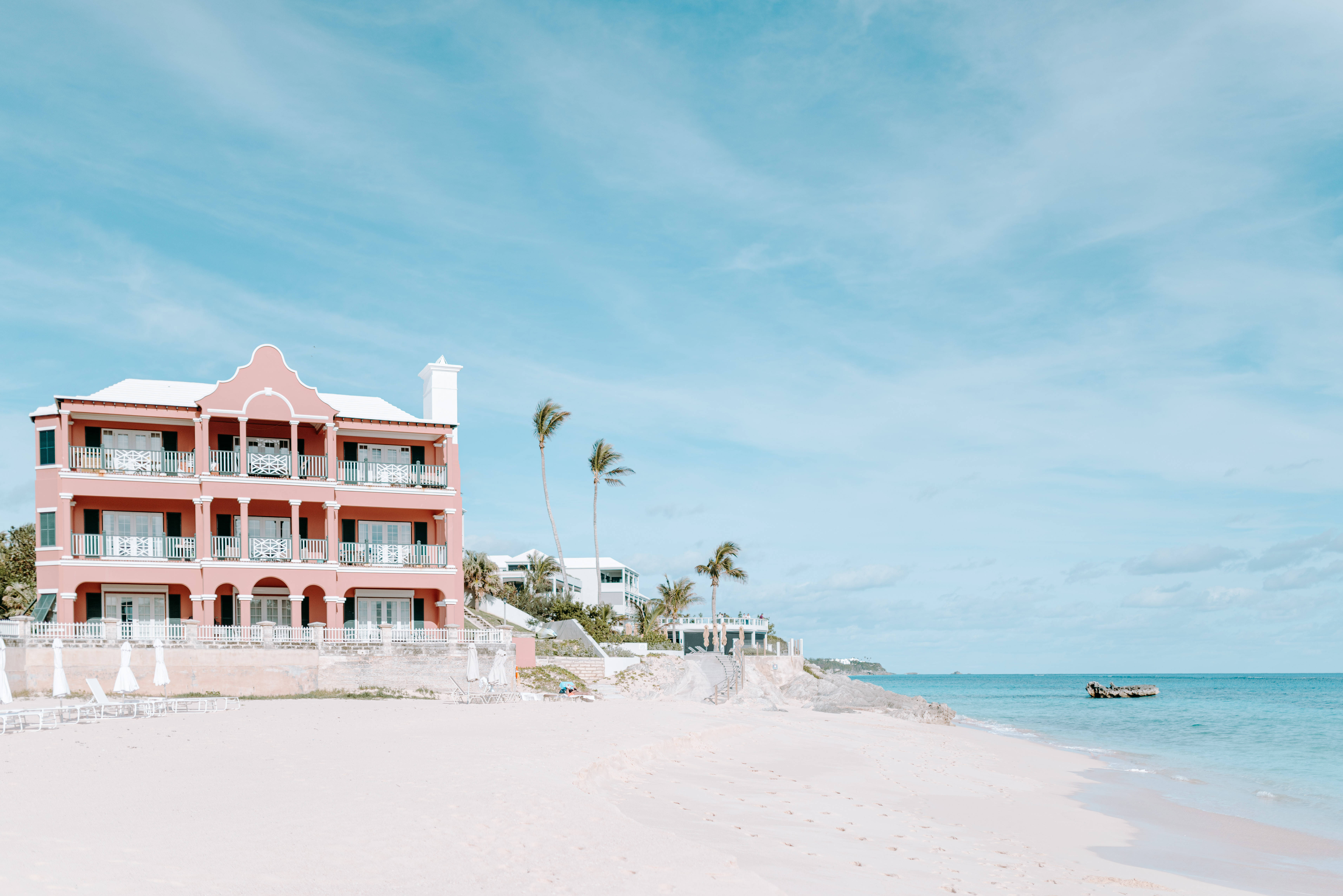 Bermuda 桌布 壁紙 — 一間粉彩粉紅色海濱飯店，設有白色陽台，矗立在沙灘上，天空湛藍，白雲點點。