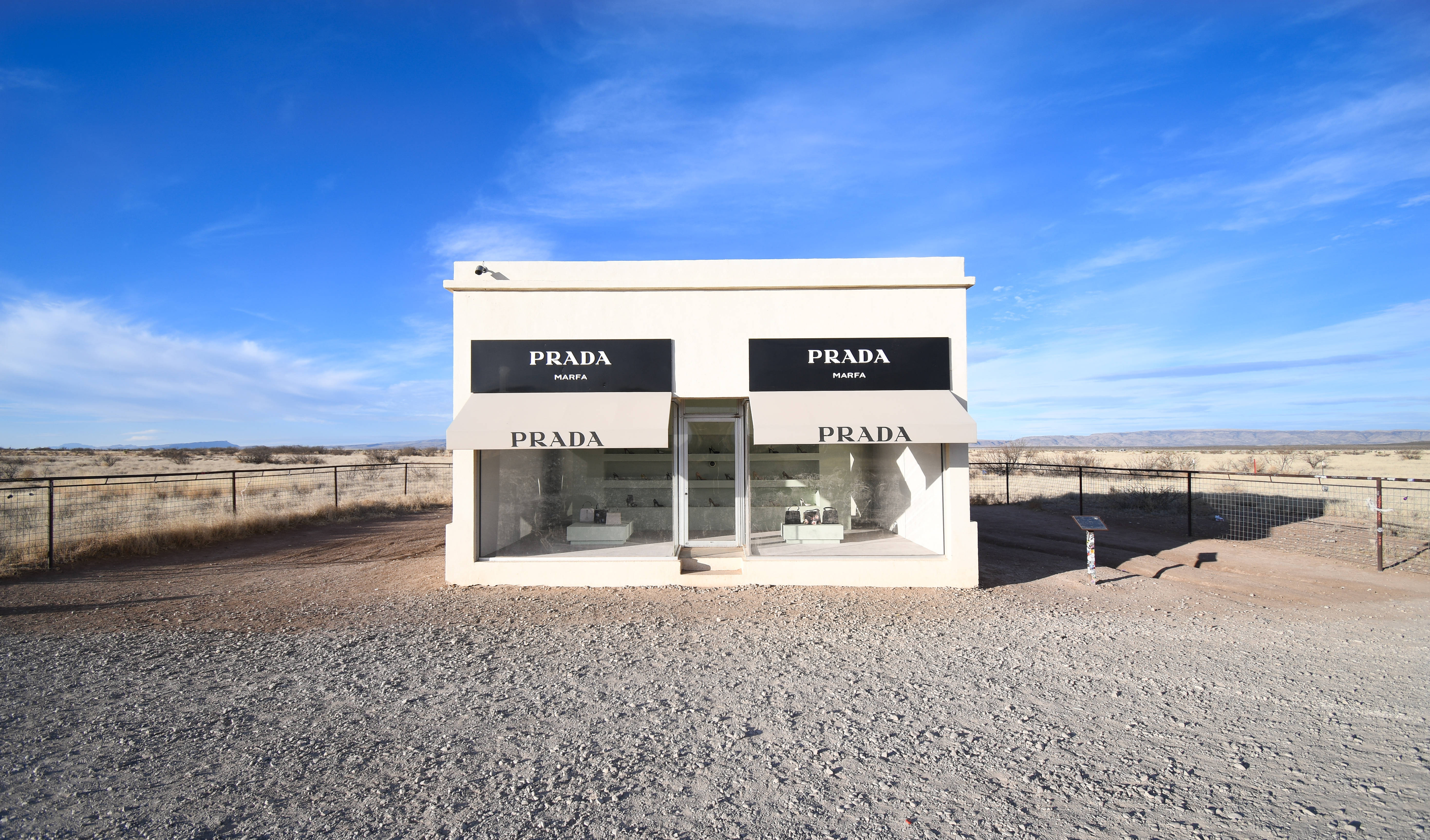Prada 桌布 壁紙 — 一間純白色的Prada精品店獨自矗立在廣闊乾旱的沙漠中，天空湛藍，雲朵縷縷。