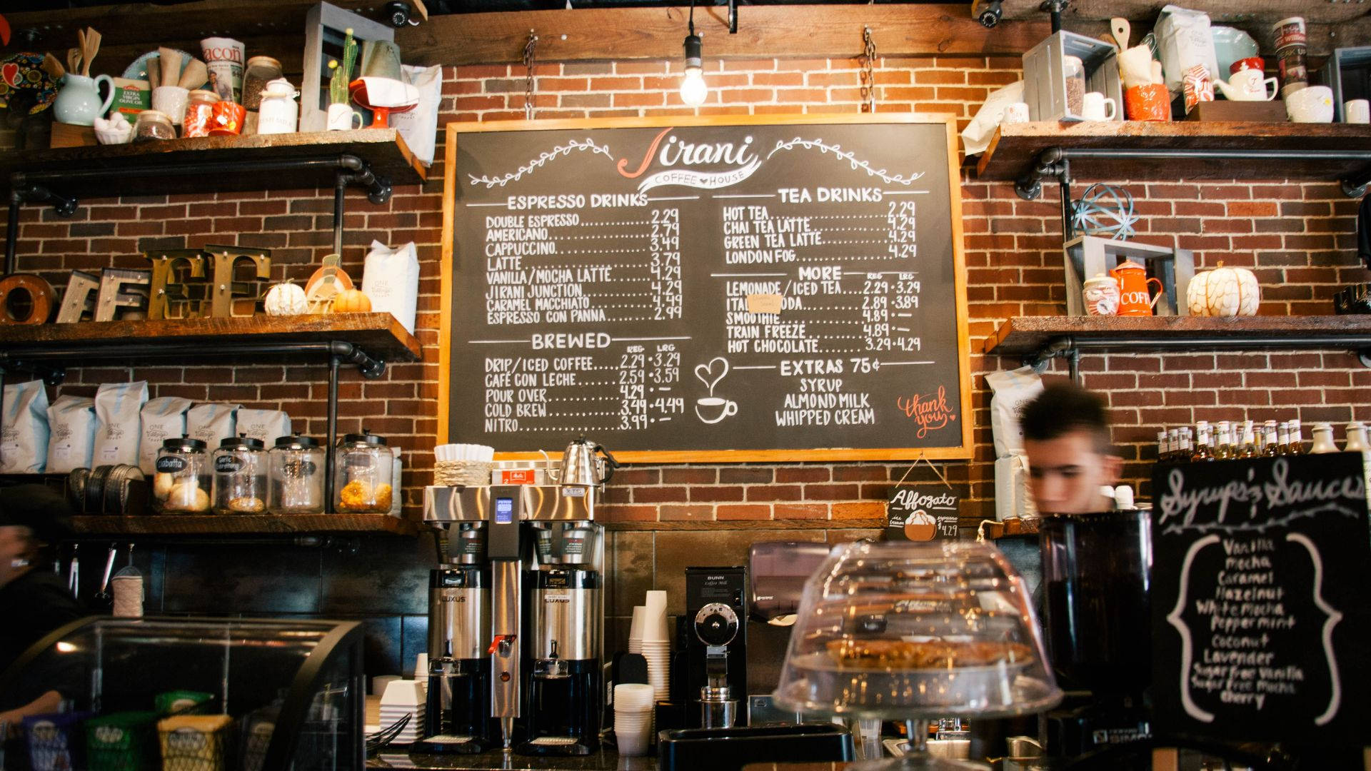 drinks 桌布 壁紙 — 一間舒適的咖啡店，設有裸露磚牆、擺滿馬克杯的架子，以及顯示濃縮咖啡和茶飲的黑板菜單。