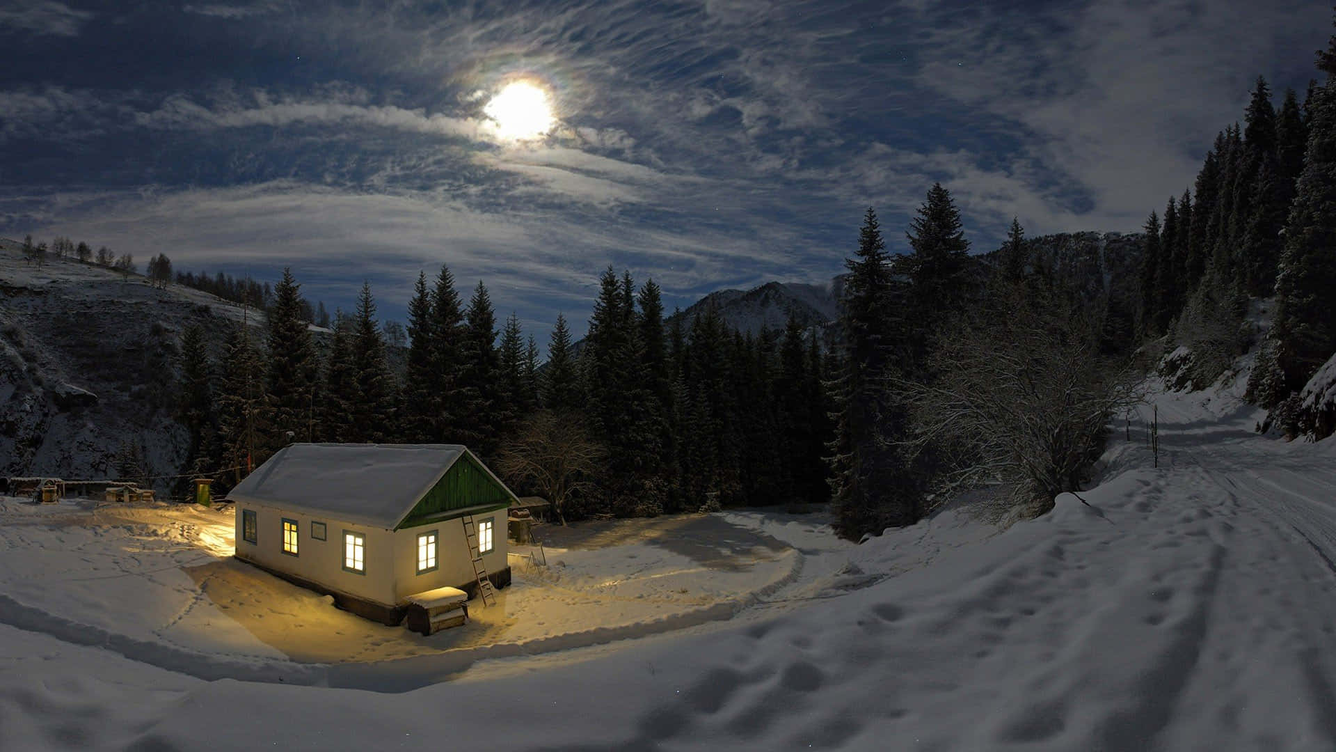 full moon 桌布 壁紙 — 一間舒適的小屋在明亮的月光下溫暖地發光，周圍環繞著白雪覆蓋的松樹和山脈。