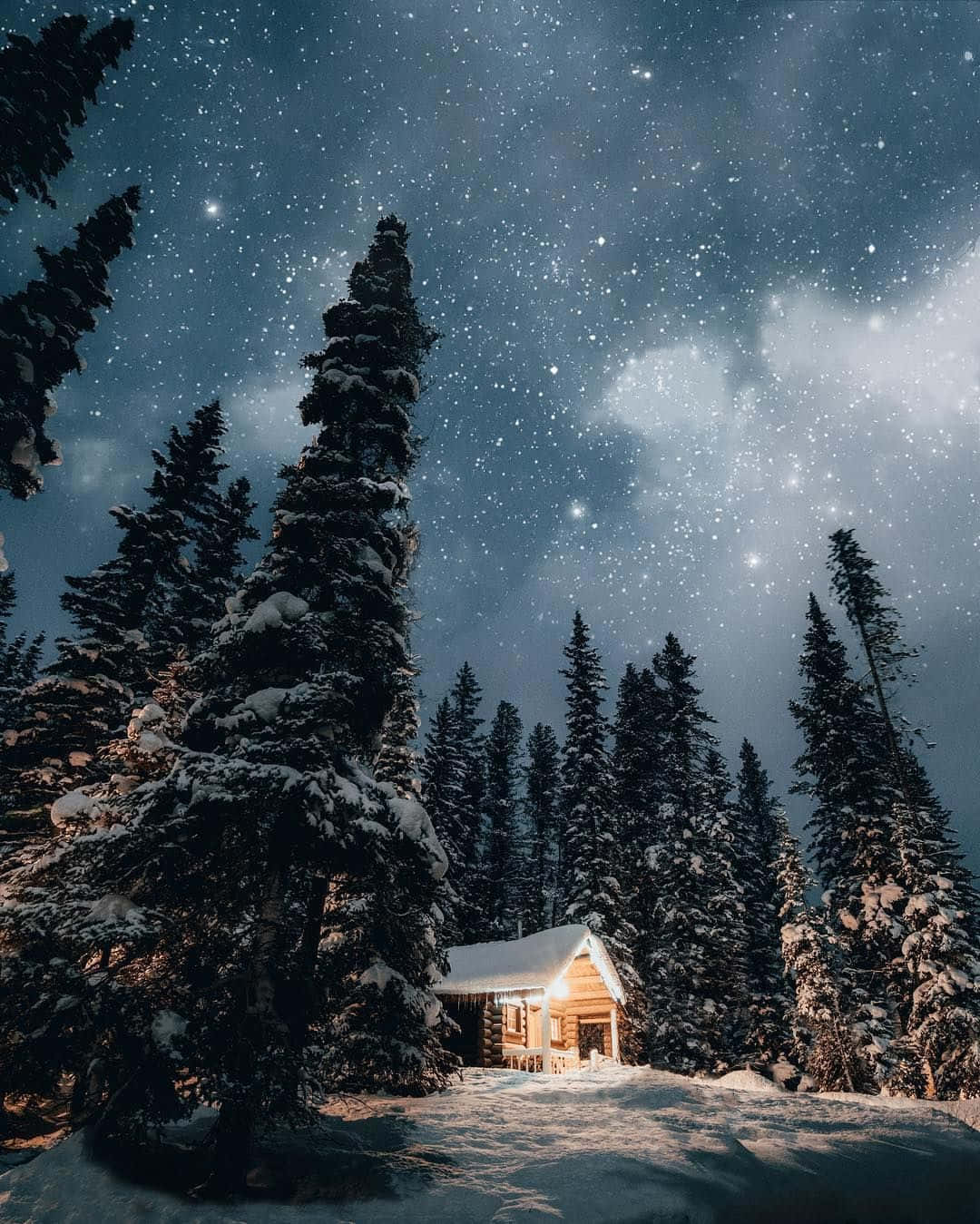 Cozy iPhone 桌布 壁紙 — 一間舒適的木屋，坐落在白雪覆蓋的松樹林中，上方是繁星點點的冬夜天空。
