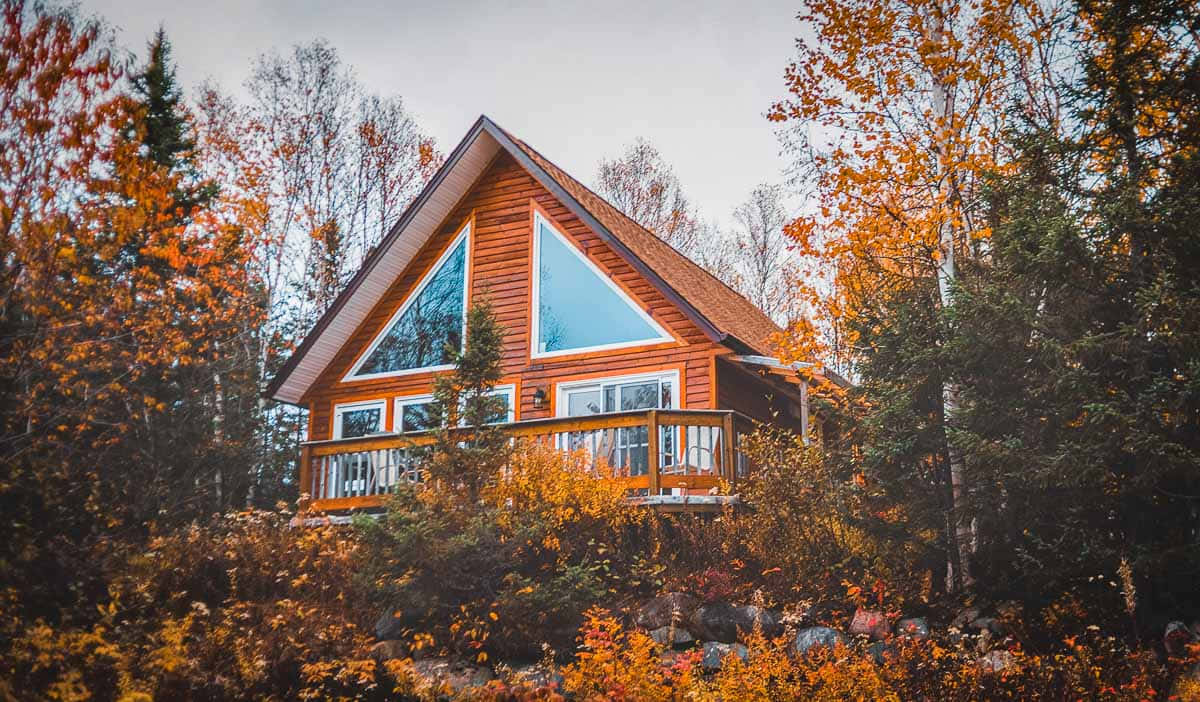 Fall Cabin 桌布 壁紙 — 一間舒適的木屋，帶有三角形窗戶，坐落在色彩繽紛的秋樹之間，天空柔和多雲。