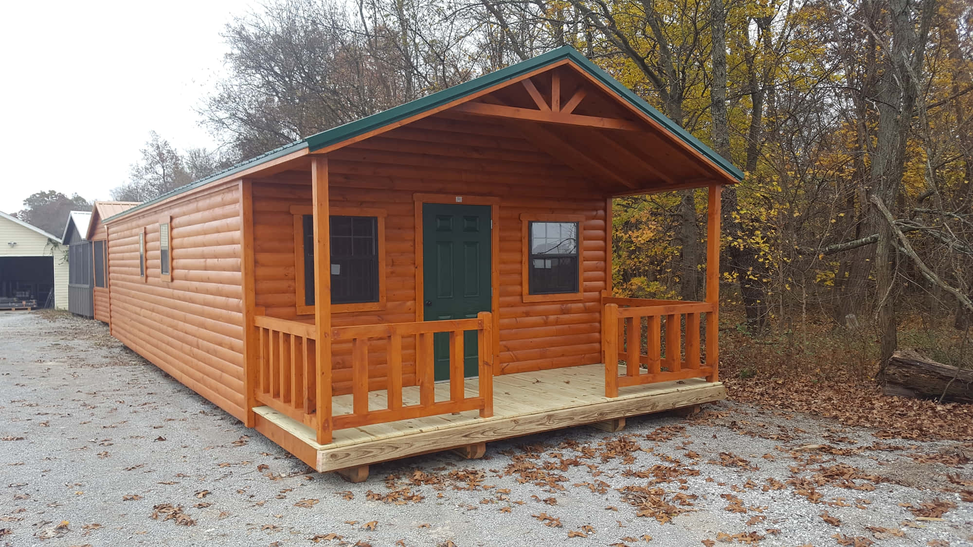 Log Cabin 桌布 壁紙 — 一間舒適的木屋，帶有綠色門和門廊，矗立在秋天的樹林中，碎石地上散落著落葉。